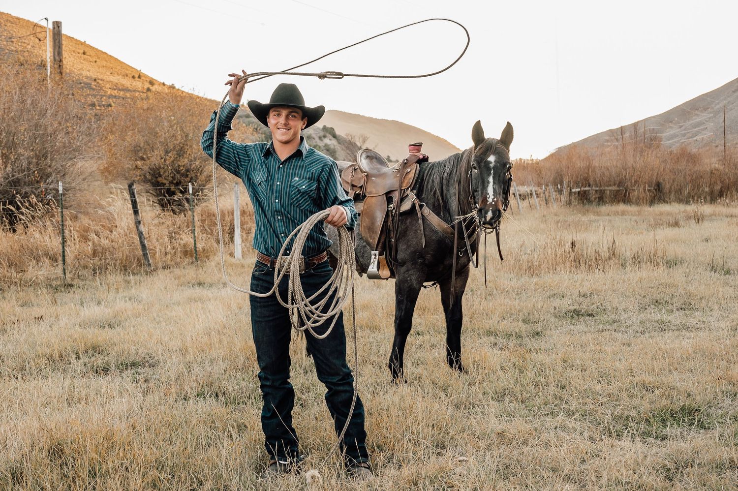 A cowboy practices with a lasso beside a dark horse in a mountain valley.