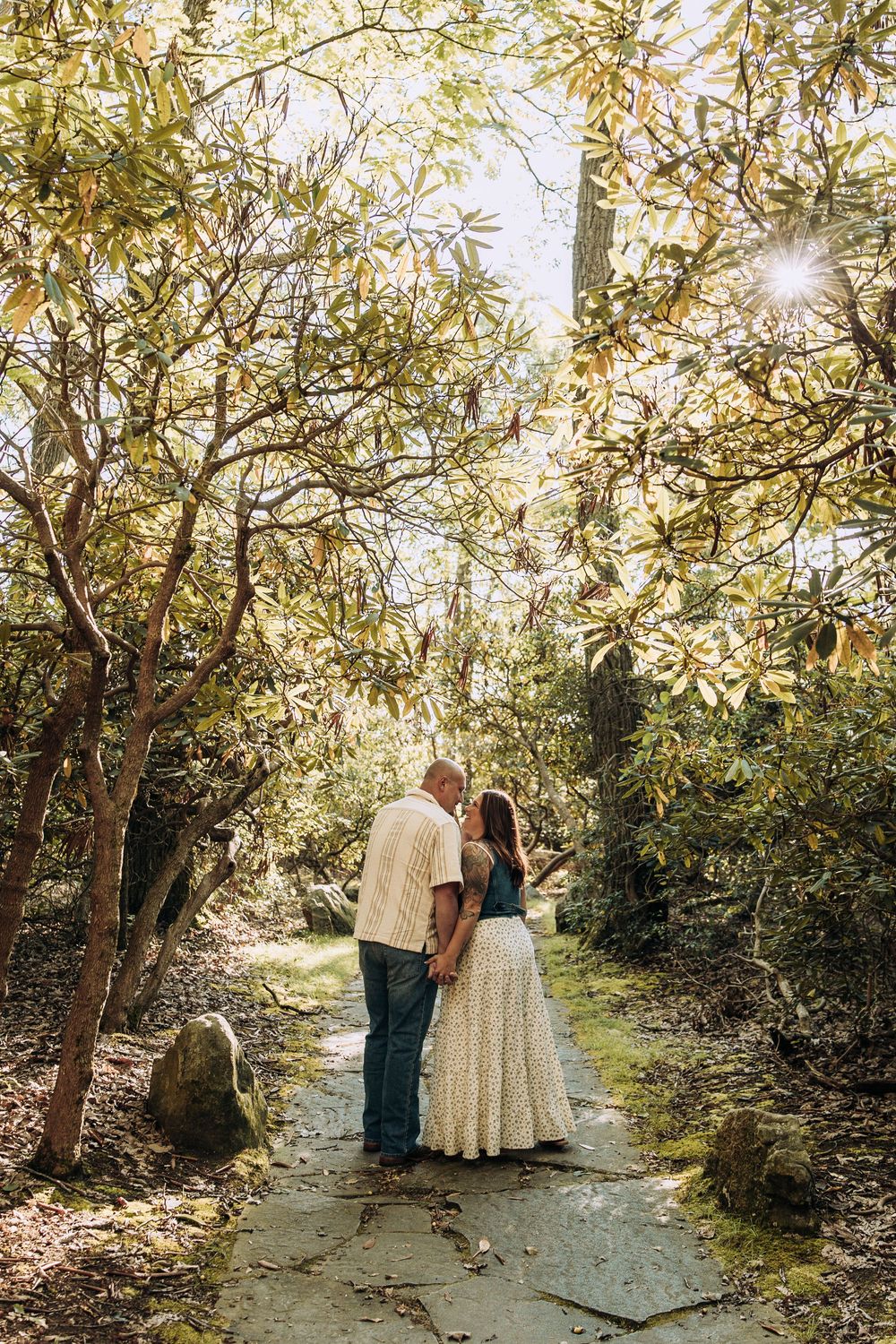 Couple holding hands and sharing a romantic moment on a stone path surrounded by sunlit trees.