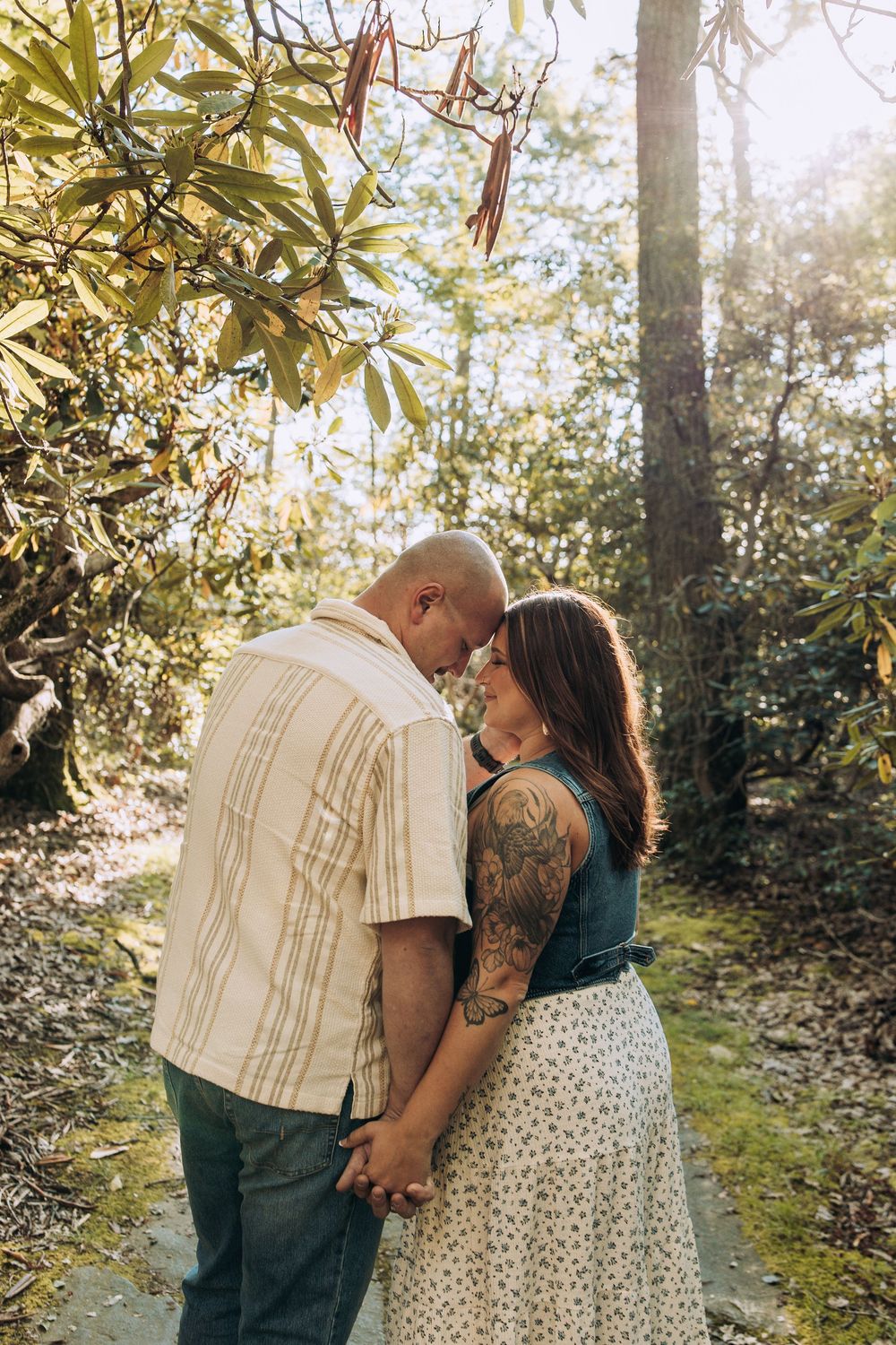 Couple with tattooed woman touching foreheads tenderly while holding hands in a sunlit forest.
