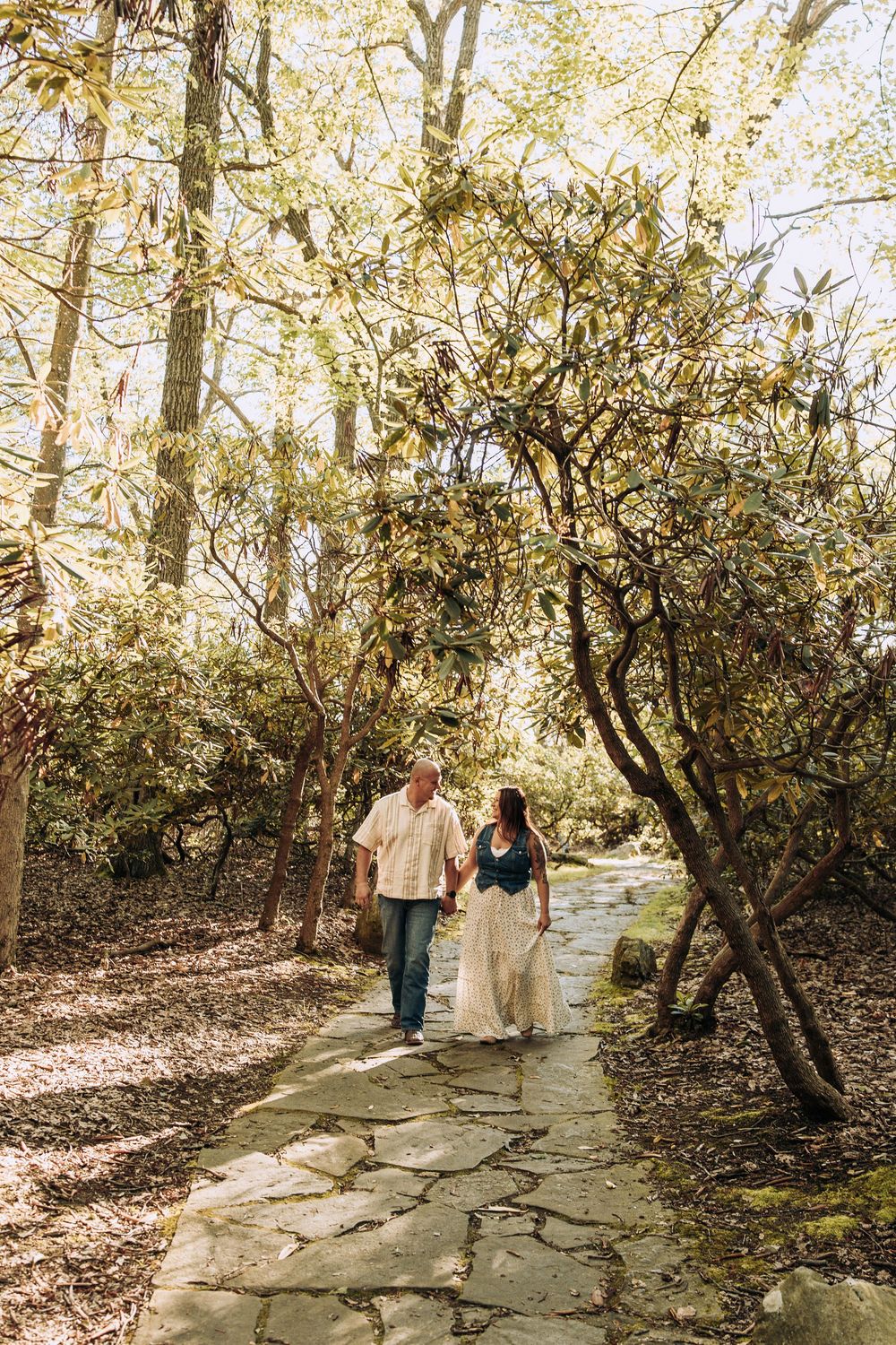 Couple walking hand in hand along a stone garden path surrounded by lush green trees.