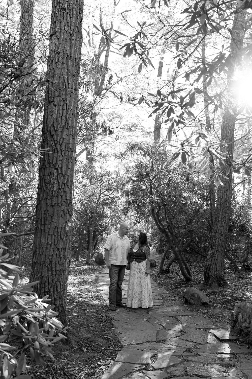 Black and white photo of a couple holding hands on a stone path surrounded by tall trees.