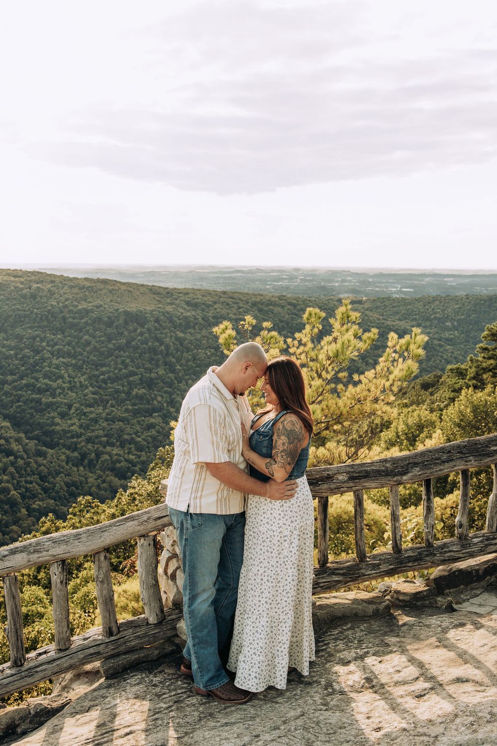 Couple embracing at a scenic mountain overlook with lush green forested valley in the background.