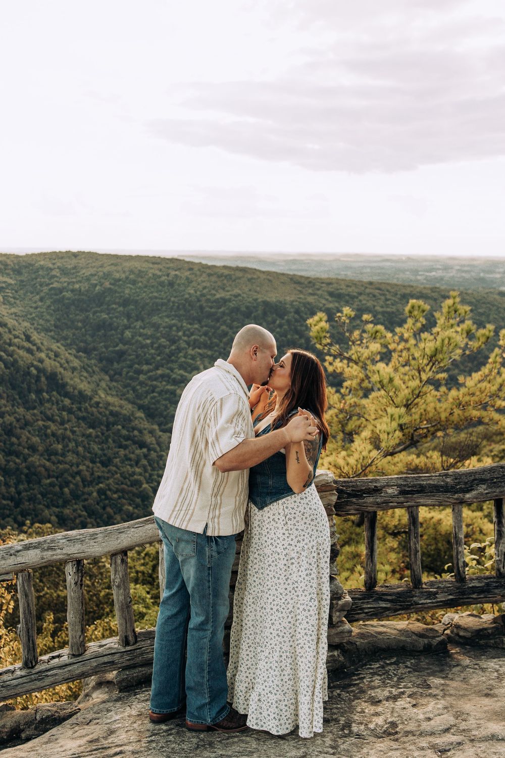 Couple sharing a romantic kiss on a scenic overlook with lush green mountain views behind them.
