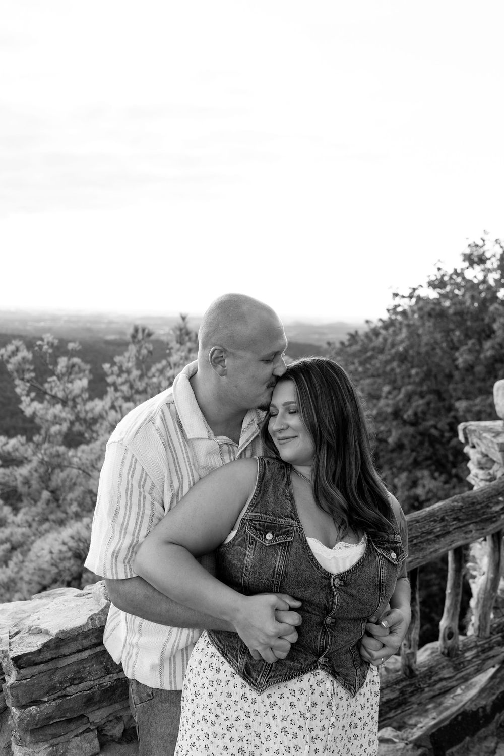 Black and white photo of a couple embracing tenderly at a stone overlook with trees in background.