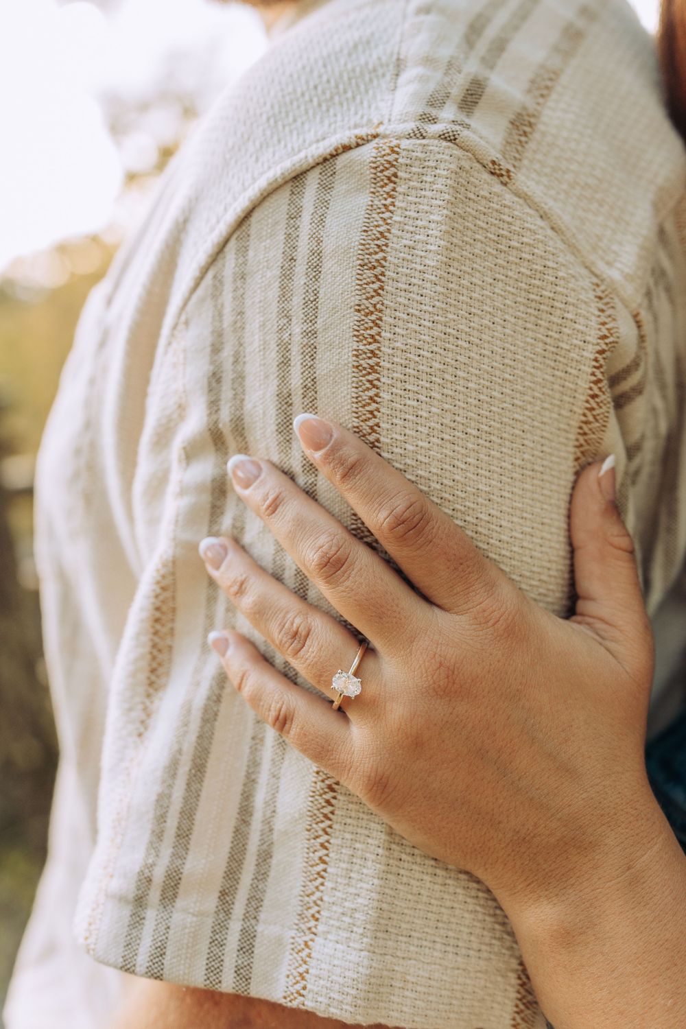 Close-up of a woman's hand with a diamond engagement ring resting on her partner's striped shirt.