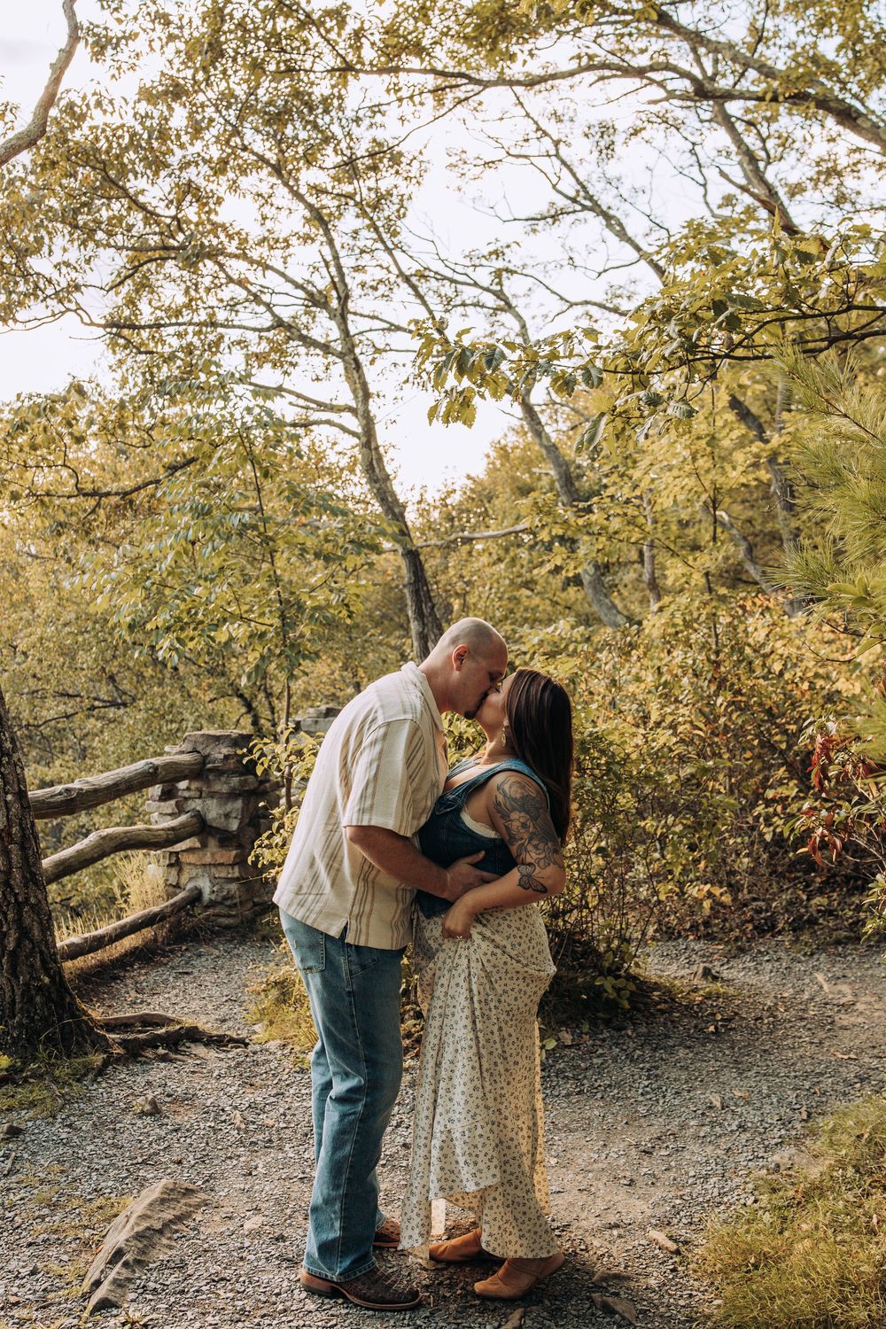 Couple sharing a kiss on a wooded trail with vibrant fall foliage and rustic fence behind them.