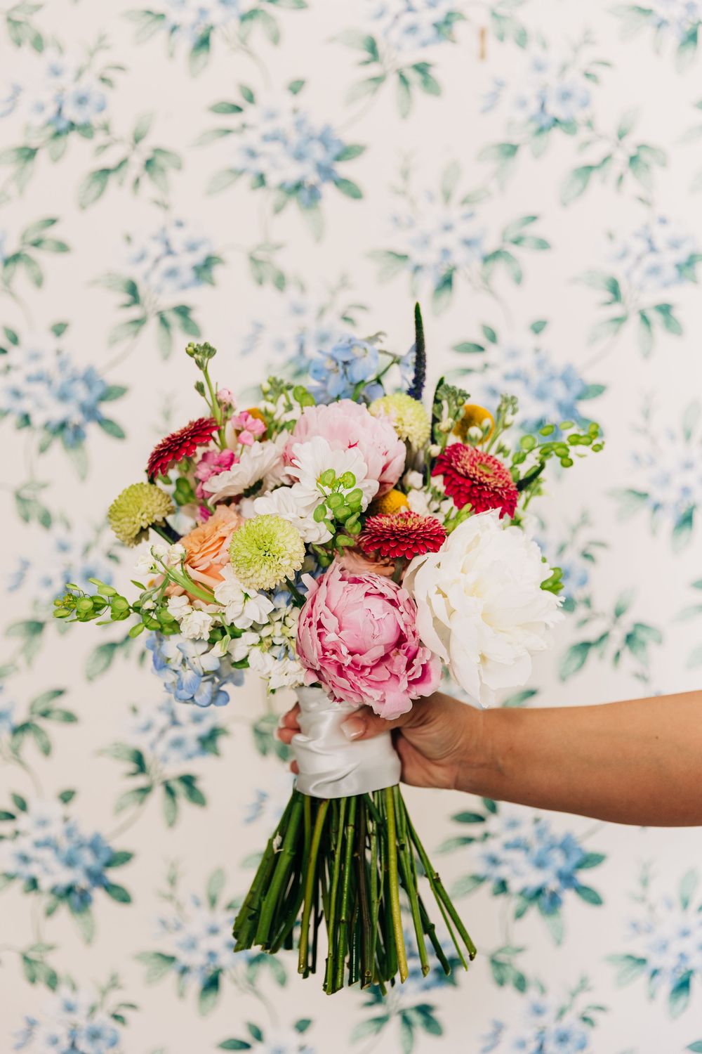 A hand holds a vibrant spring bouquet with pink peonies, white dahlias, and green accents against a floral wallpaper at The Wild Flower 301