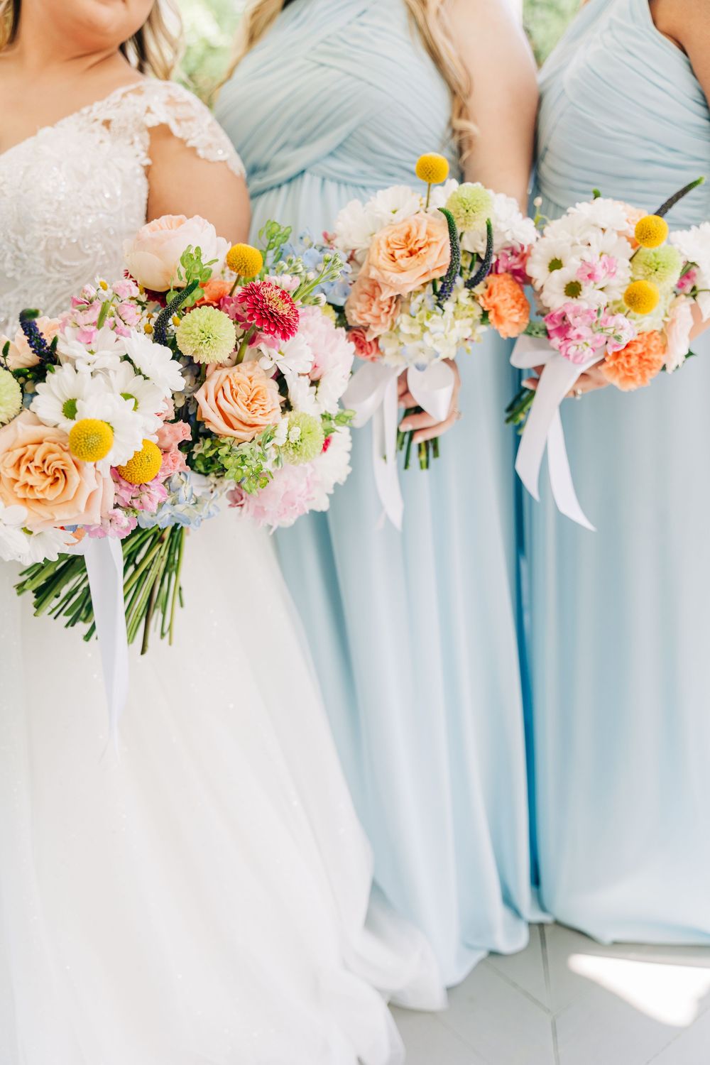 Wedding party members in light blue dresses hold vibrant coral, pink and yellow floral bouquets at The Wild Flower 301