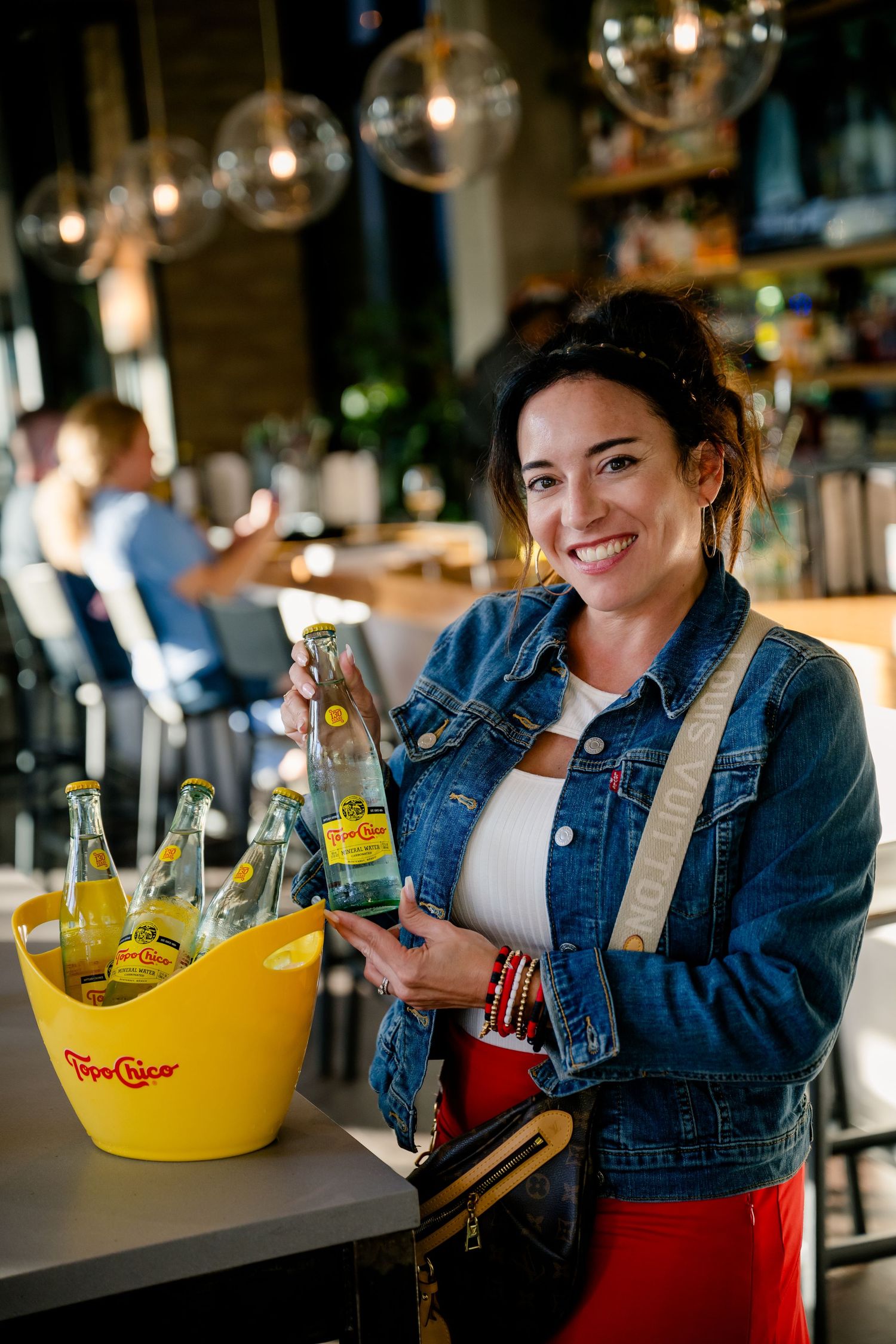 A person in denim jacket stands at a bar counter with a yellow drink bowl and bottles in a warmly lit setting.