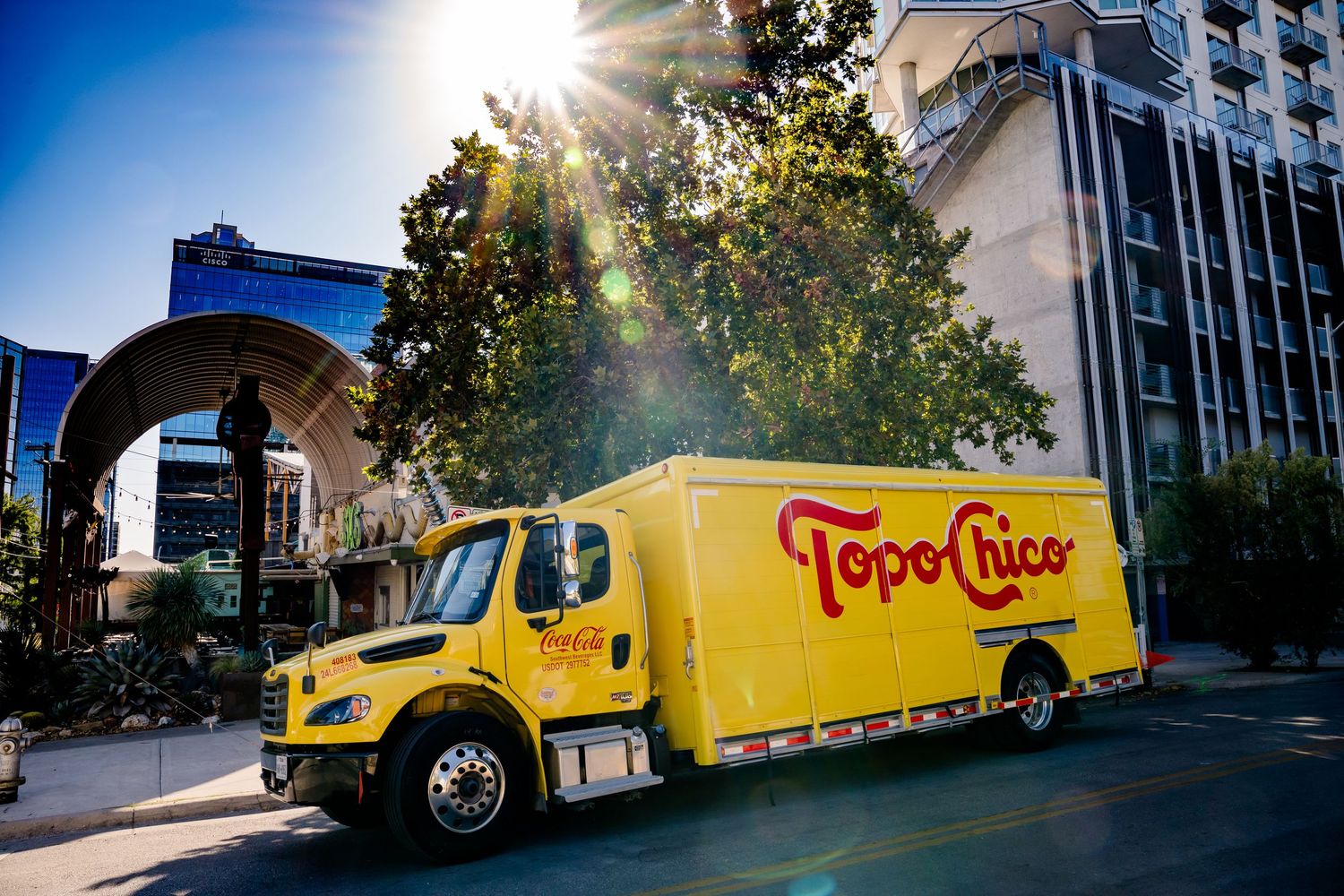 A yellow Topo Chico delivery truck parked on a sunny street with buildings and trees in the background.