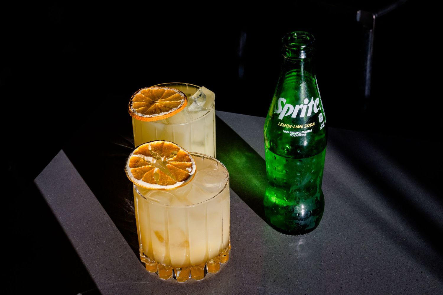 Elegant cocktail garnished with dried citrus wheel served alongside green glass bottle on dark surface.