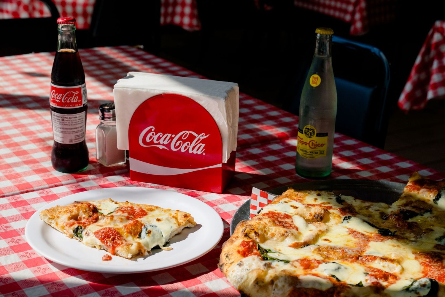 Delicious pizza slices on red and white checkered tablecloth with Coca-Cola bottles and drinks in the background.