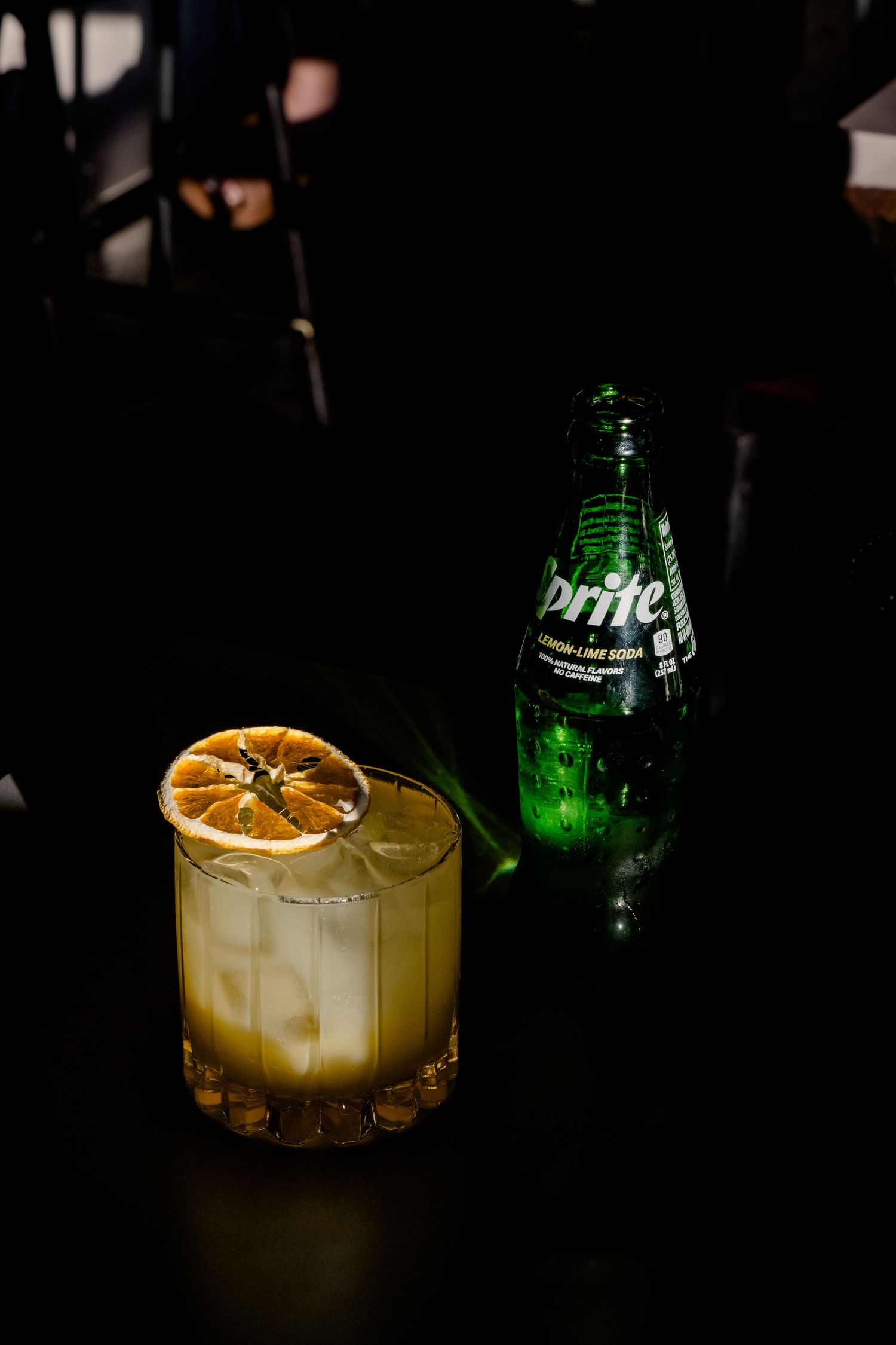 A cocktail glass with ice and dried orange garnish sits next to a green Sprite bottle on a dark surface.