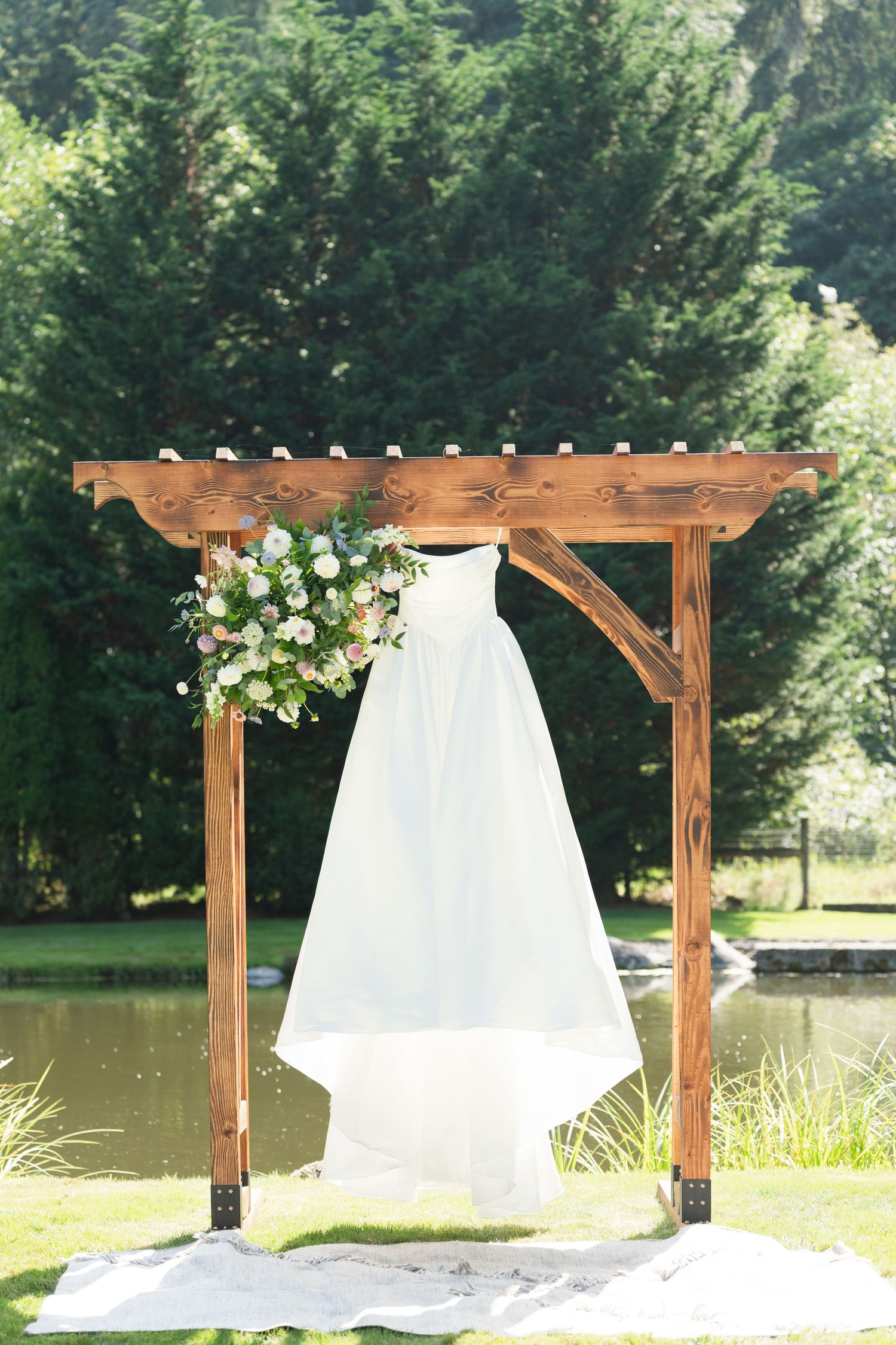 Wooden wedding pergola with flowing white fabric and floral decorations stands elegantly beside a serene pond.