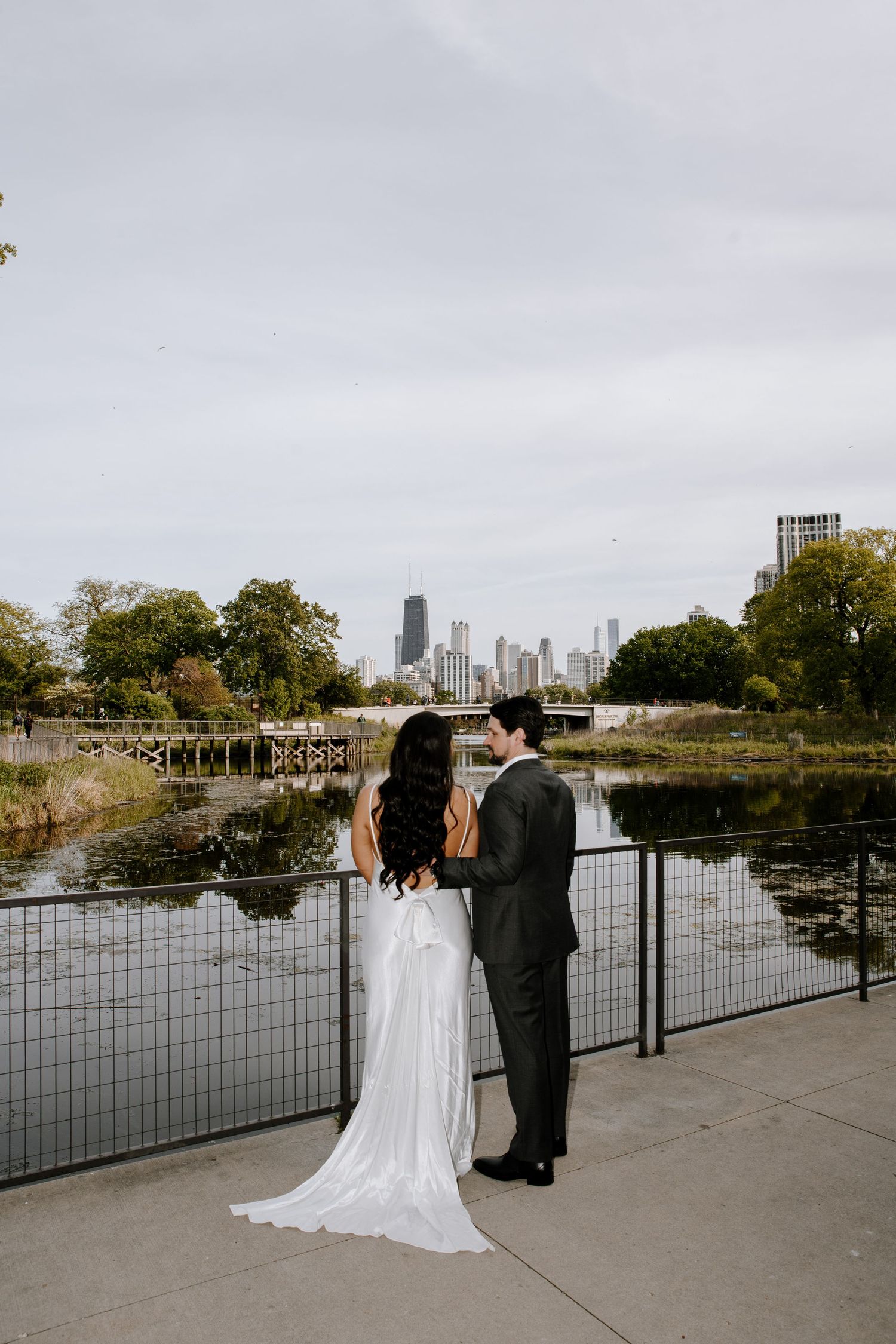 A couple in wedding attire stands at Lincoln Park's South Pond overlooking the Chicago skyline on an autumn day.