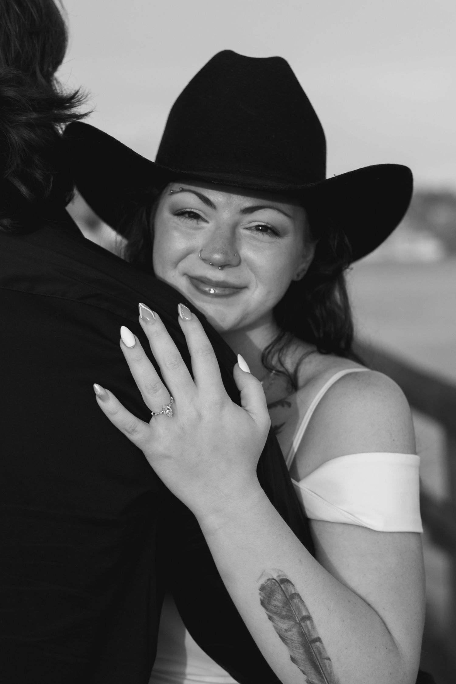 Black and white portrait of someone in a cowboy hat and white top embracing another person while showing an engagement ring.