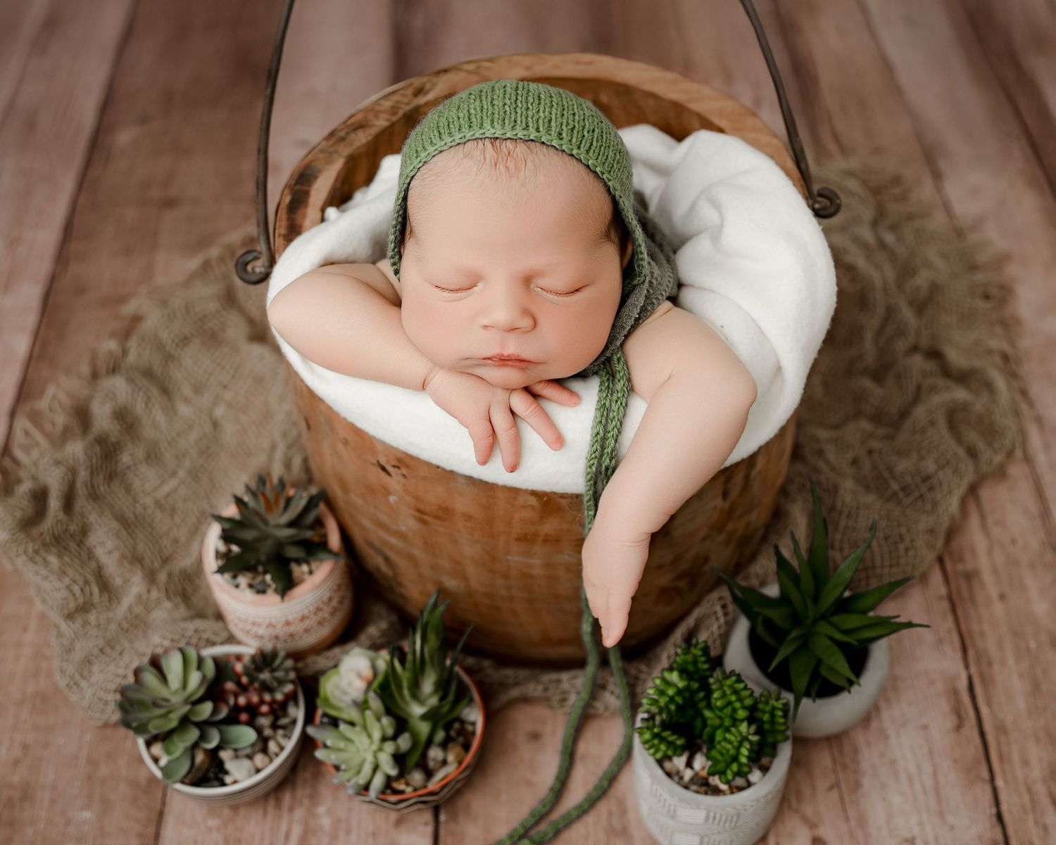 Sleeping baby in green knit hat lies peacefully in wooden bowl surrounded by small potted succulents on wooden surface.
