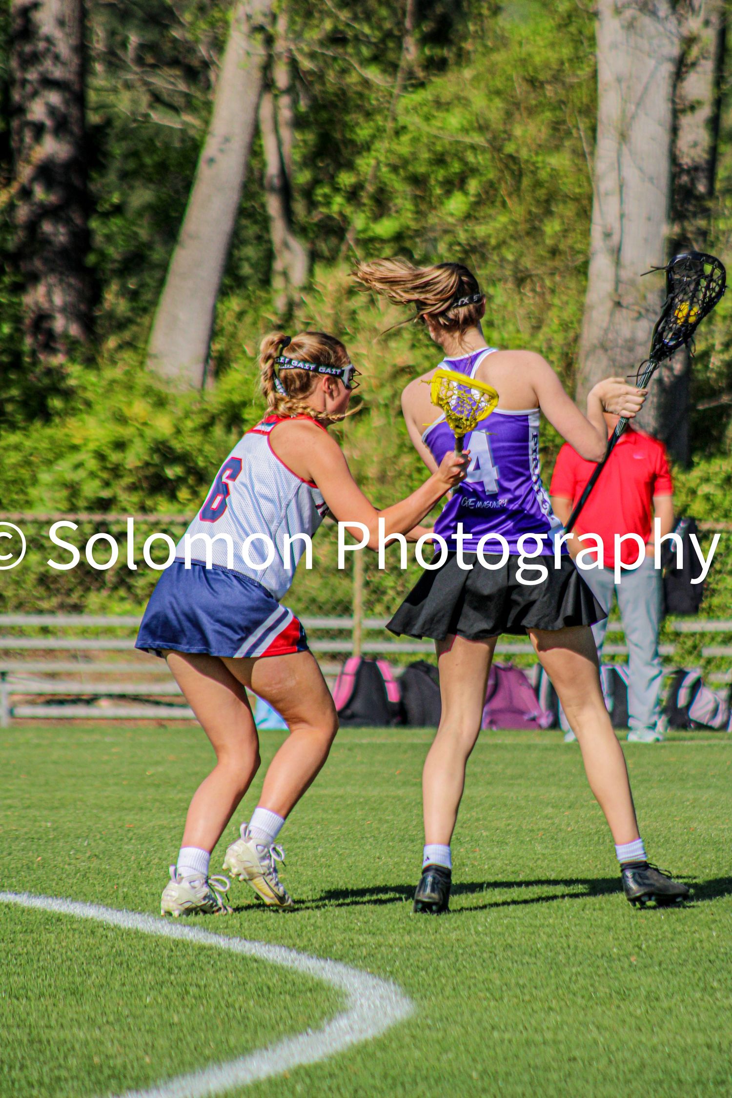 Two girls lacrosse players competing one in red white blue uniform and one in purple during game.