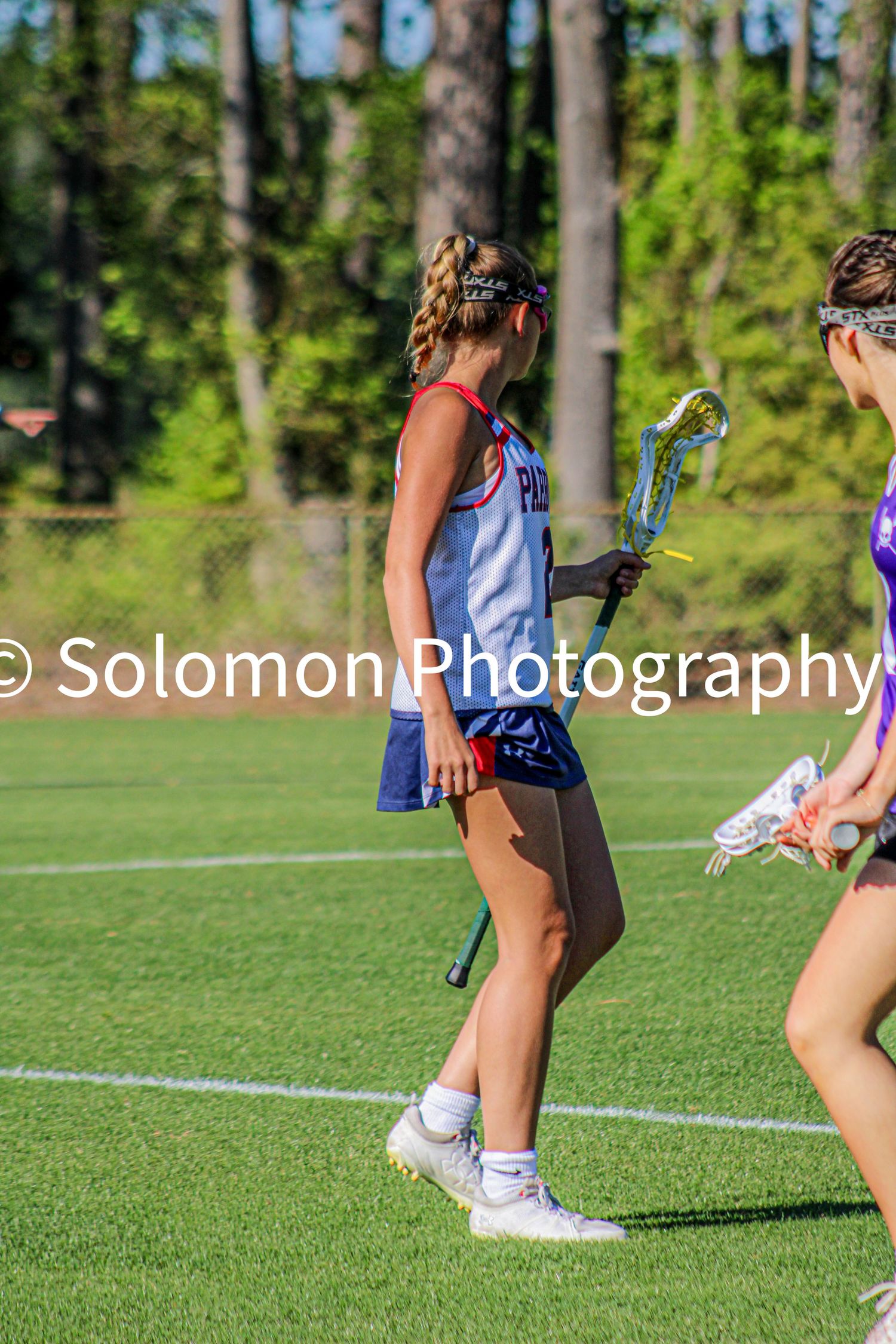Young female lacrosse player in red white and blue uniform holding stick while defending on field.