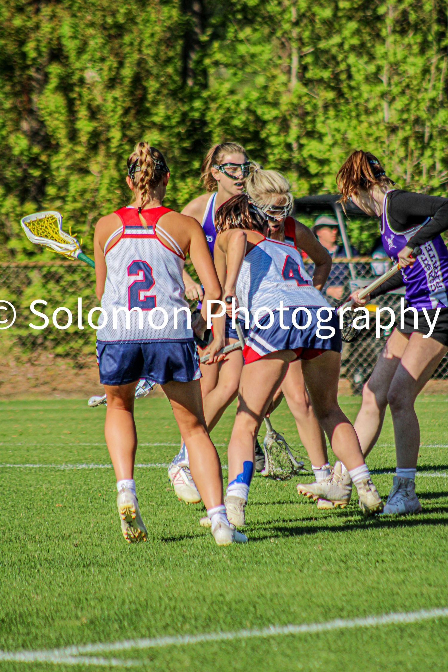 Girls lacrosse players in red white blue uniforms competing in a cluster during outdoor match.