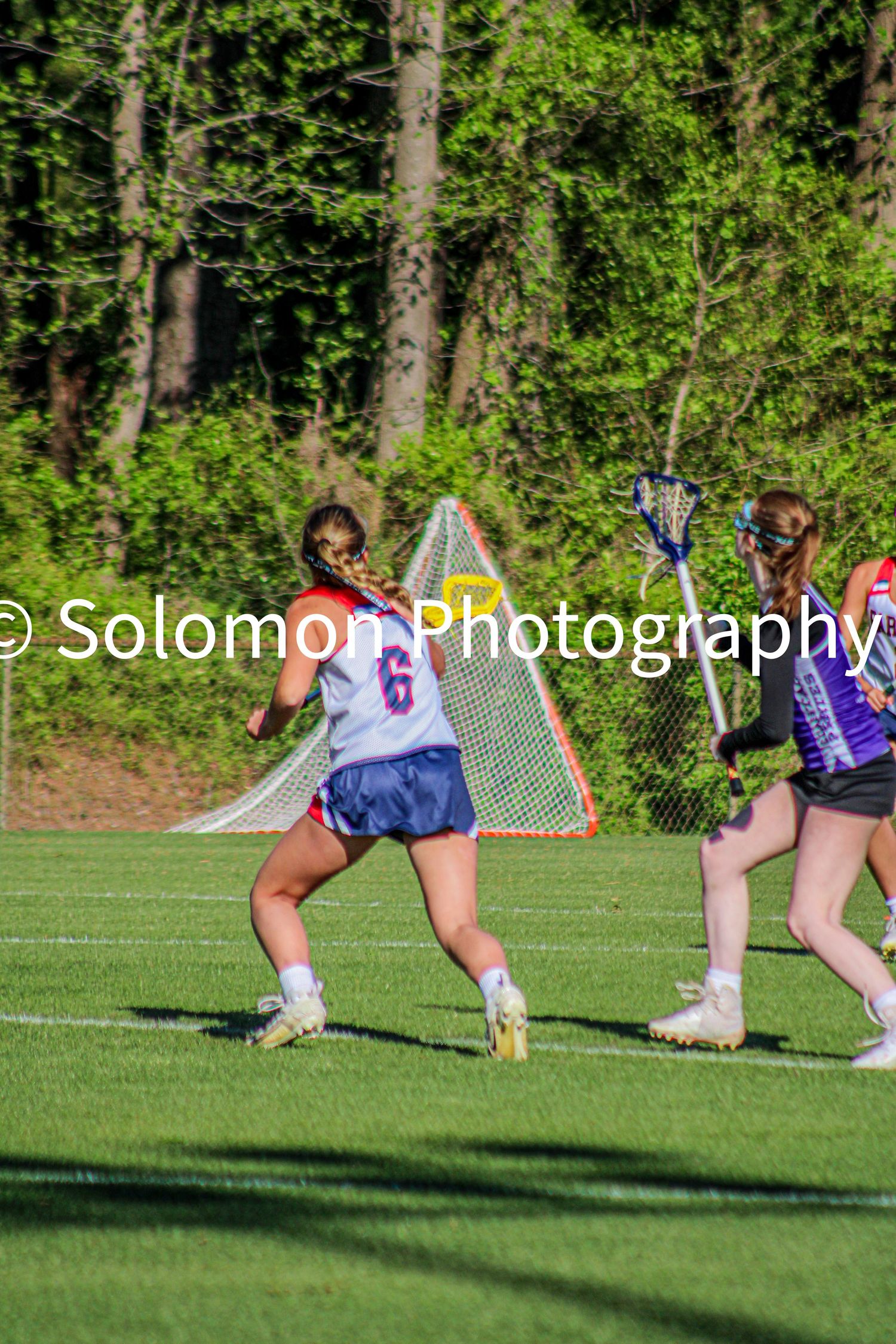 Female lacrosse player number 6 in white uniform defending against opponent near goal on green field.