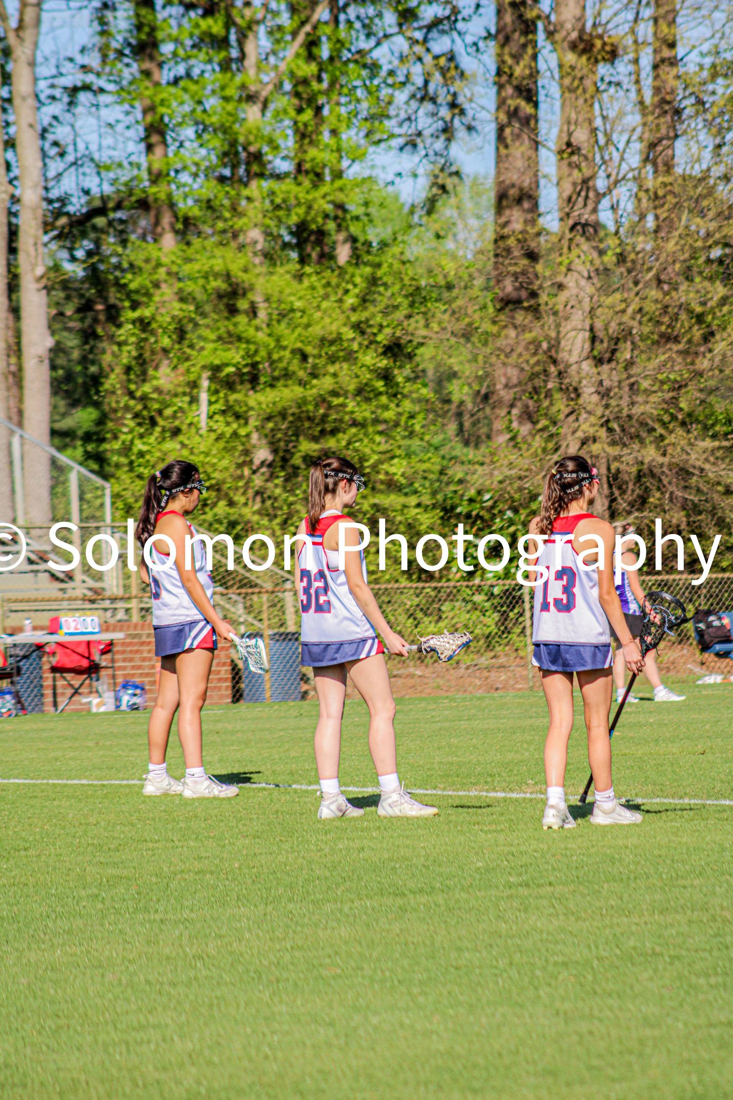 Three girls lacrosse players in white and purple uniforms standing on green field during game.
