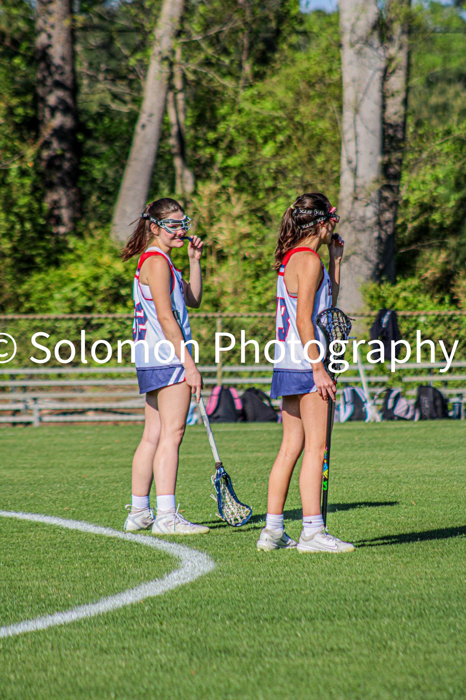 Two female lacrosse players in red white and blue uniforms talking on corner of green field.
