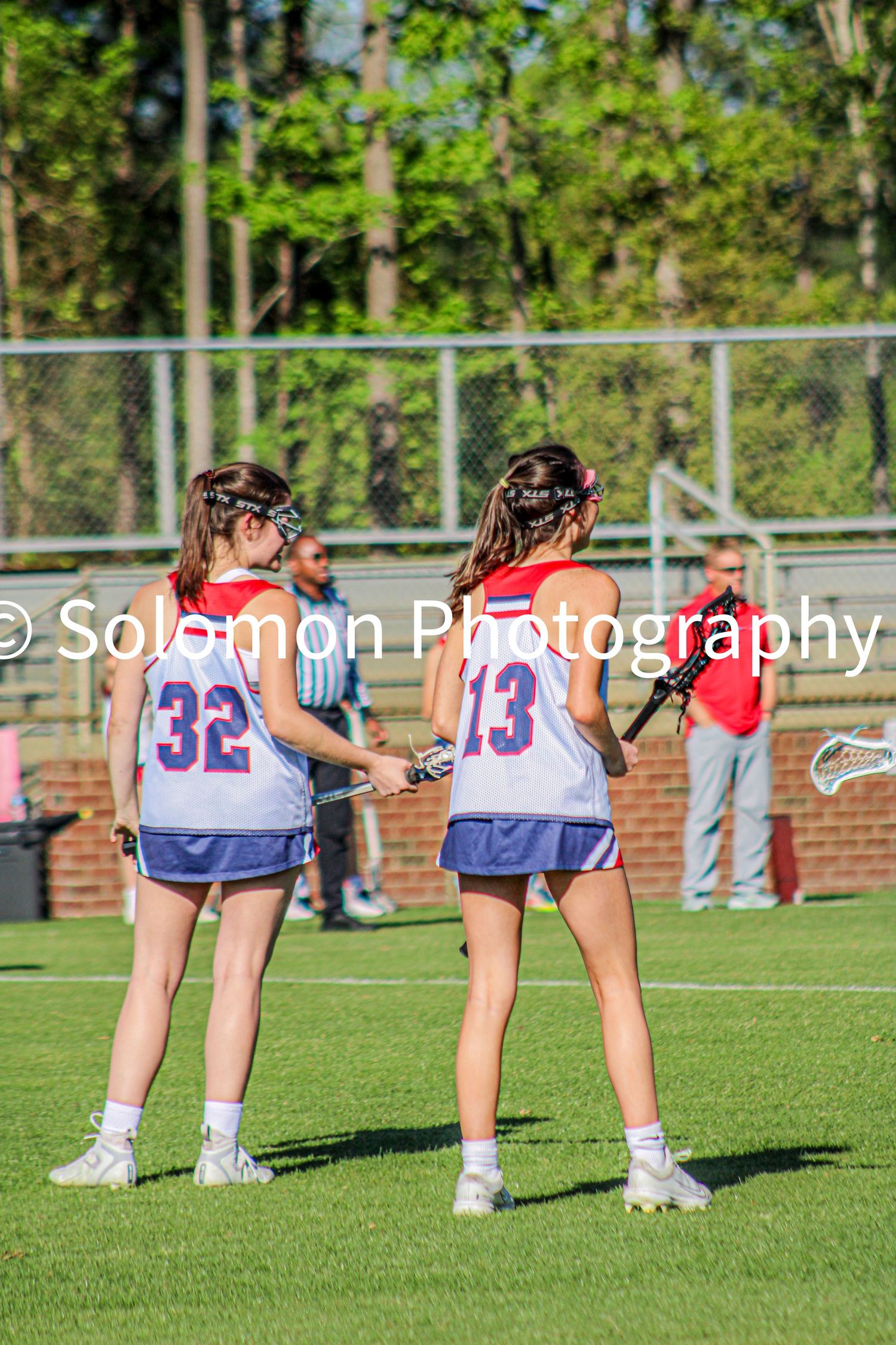 Two girls lacrosse players numbers 32 and 13 in white uniforms standing on sunny green field.