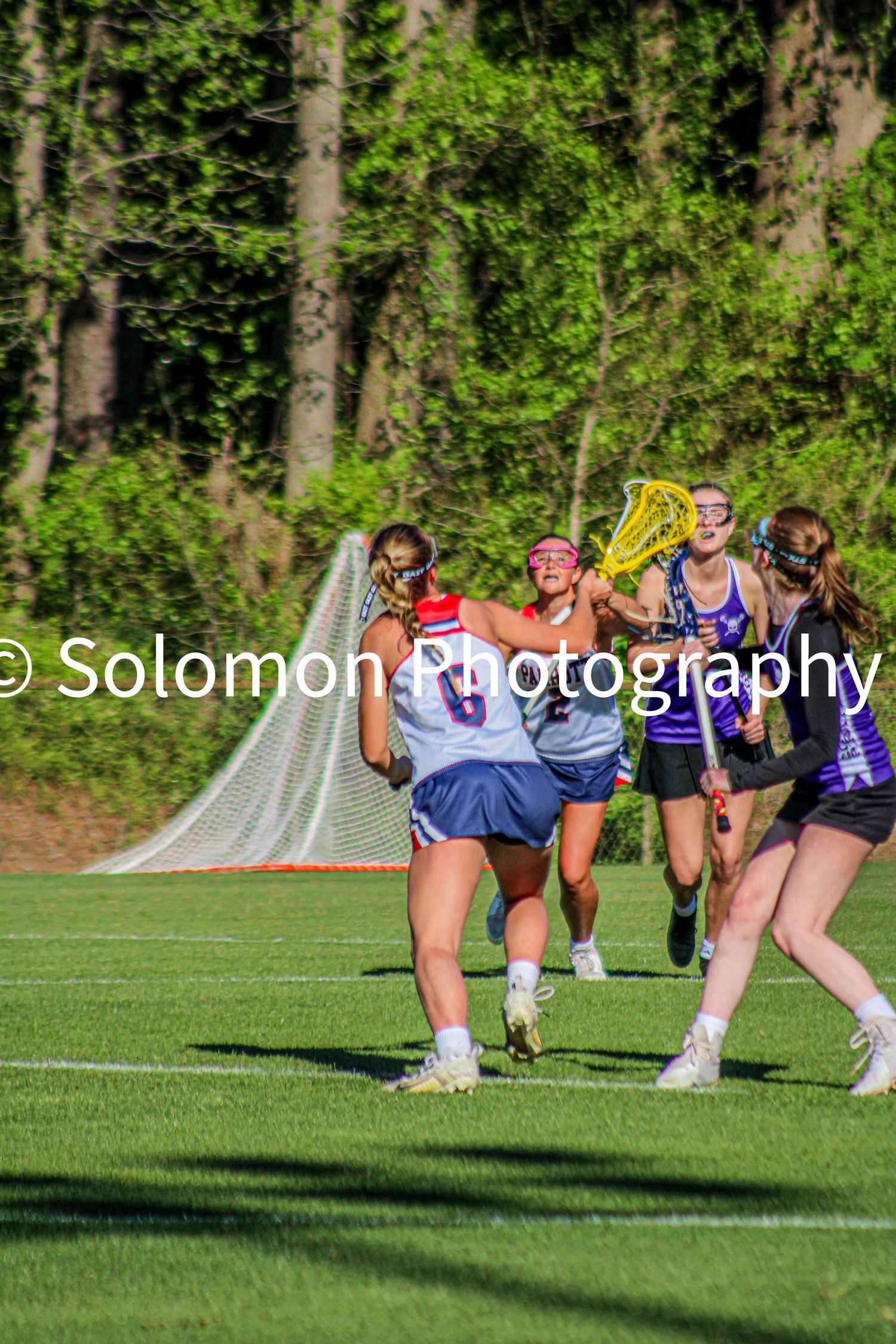 Girls lacrosse players in white and purple uniforms competing near goal during outdoor game.