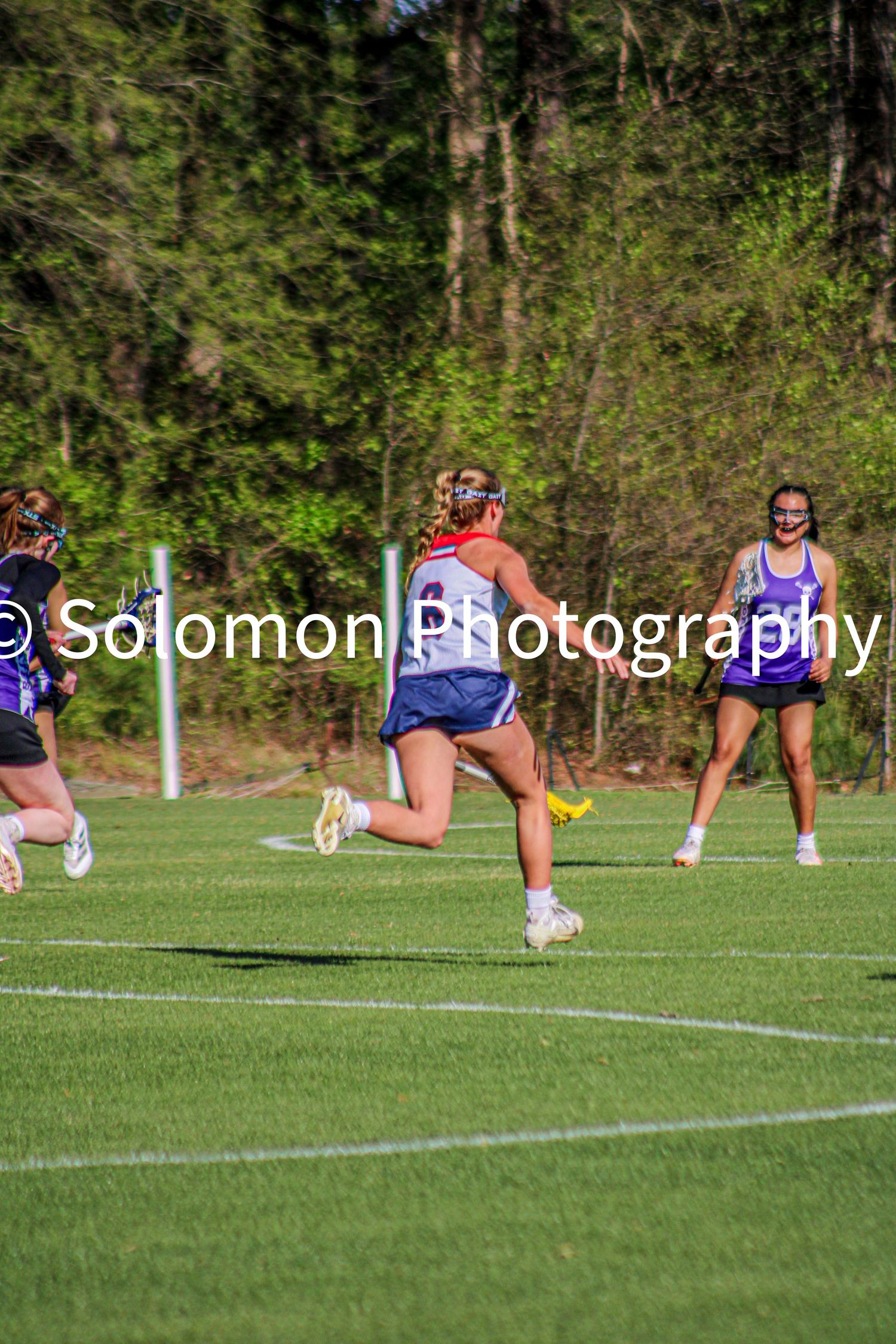 Girls lacrosse players in purple and white uniforms compete on a green turf field during a sunny outdoor game.