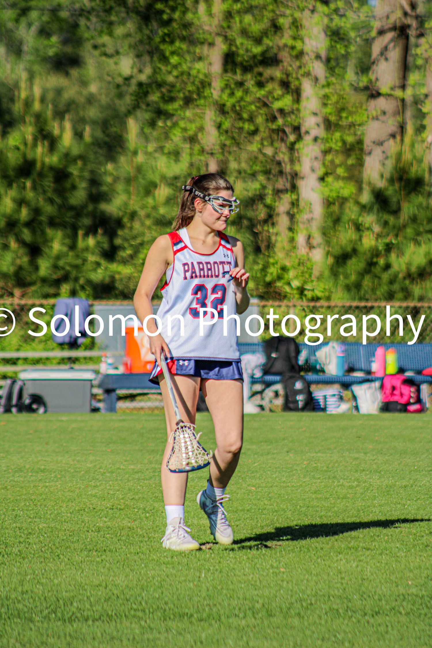 Female lacrosse player in Parrots number 33 jersey holds a stick on a green field in bright sunlight.