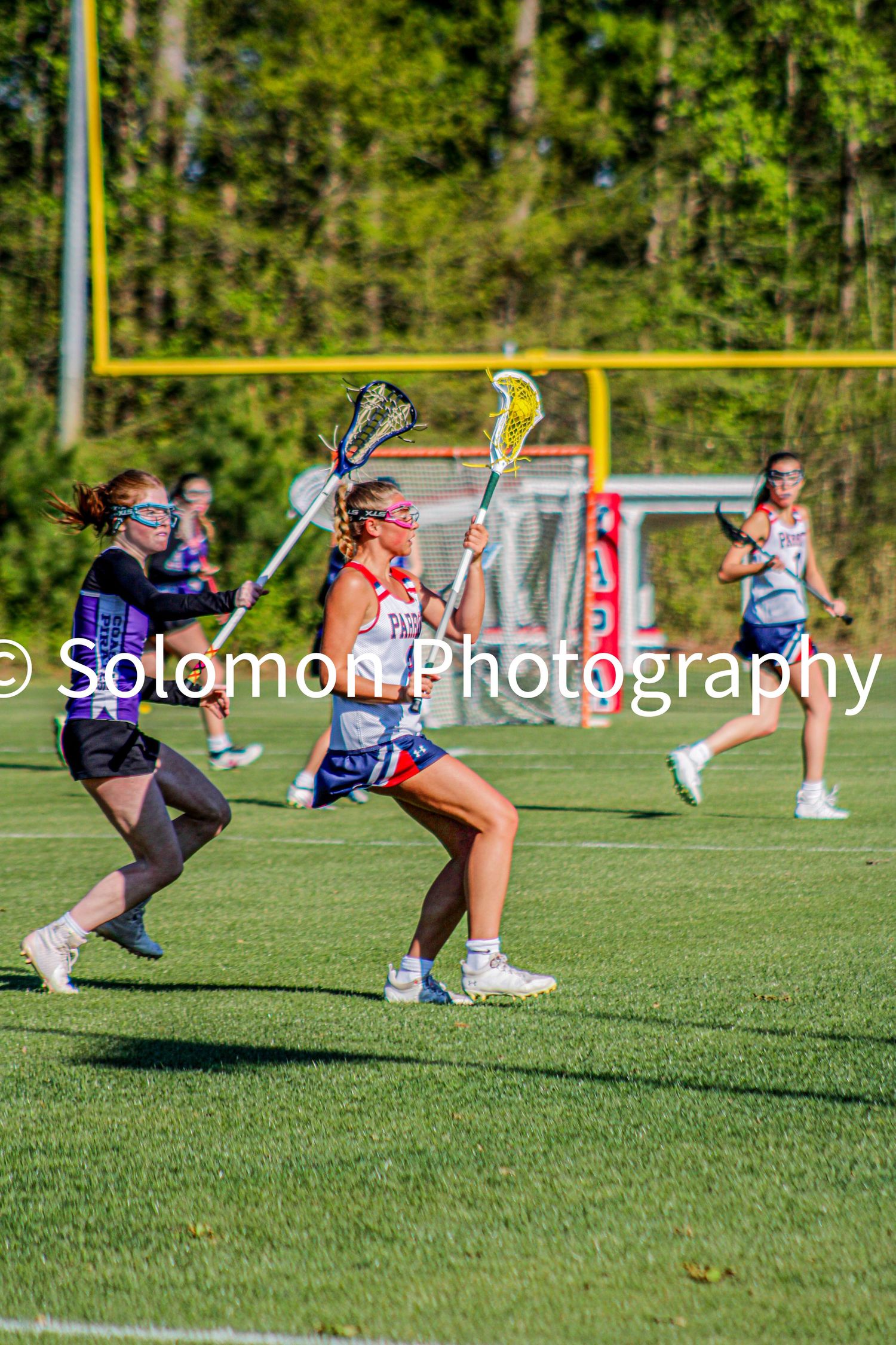 Girls lacrosse player in red white and blue uniform cradling ball while being defended during outdoor game.