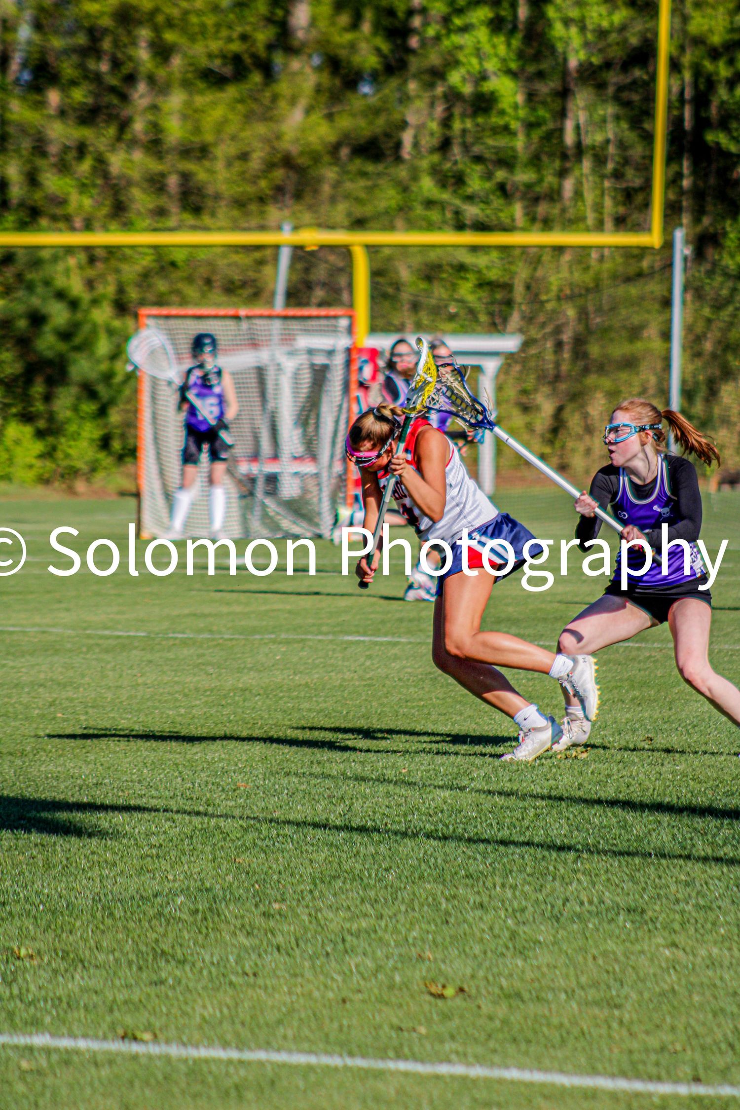 Girls lacrosse players competing on turf field during sunny game.