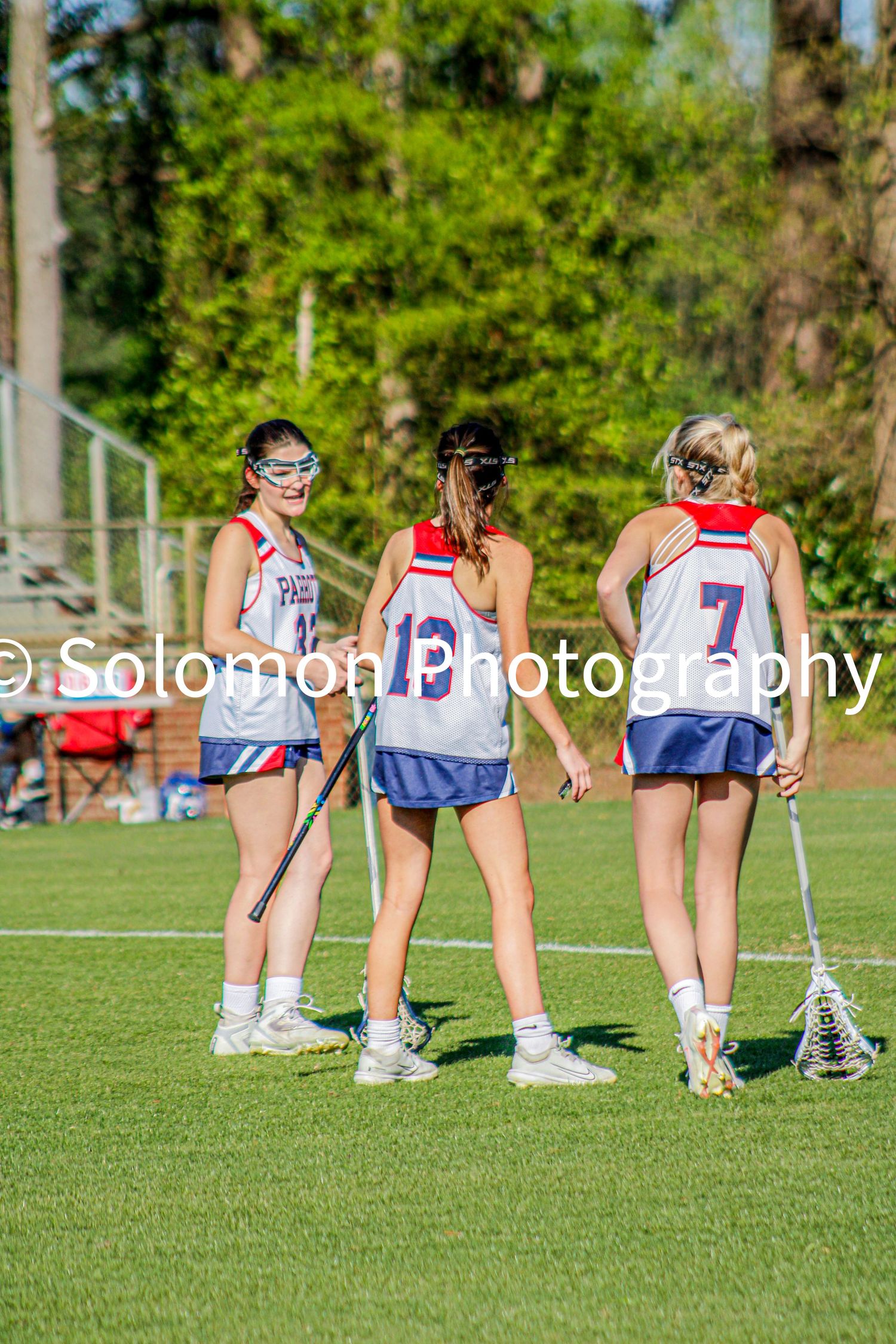 Three girls lacrosse players in red white and blue uniforms standing together on green field.
