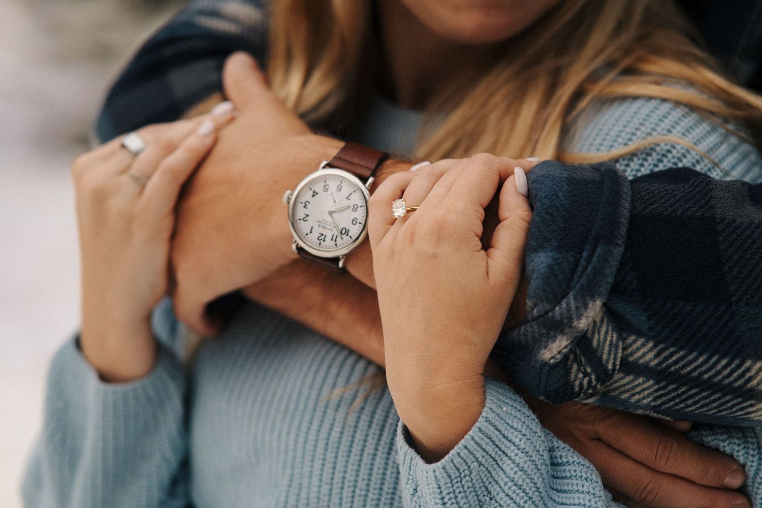 Close-up of hands wearing silver watch and rings against a blue knit sweater and dark plaid scarf.