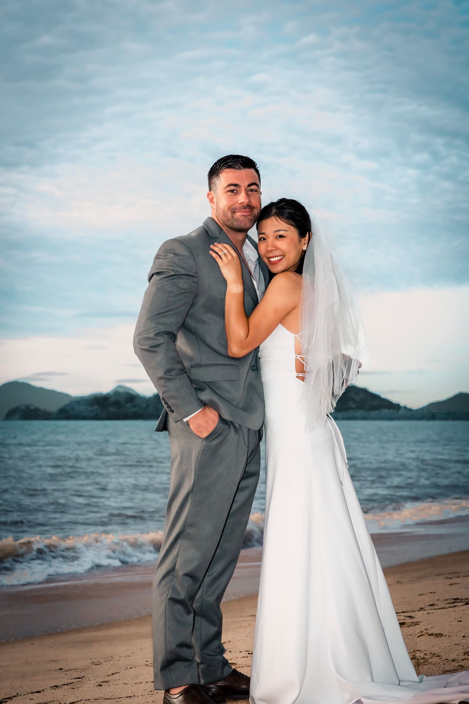 A newlywed couple embraces on a beach at sunset with mountains and ocean waves in the background.