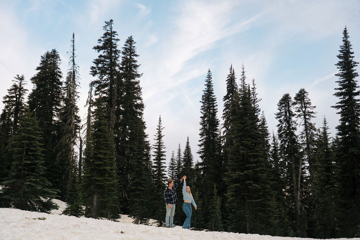 A couple stands together on a snowy slope surrounded by tall evergreen trees under a winter sky.