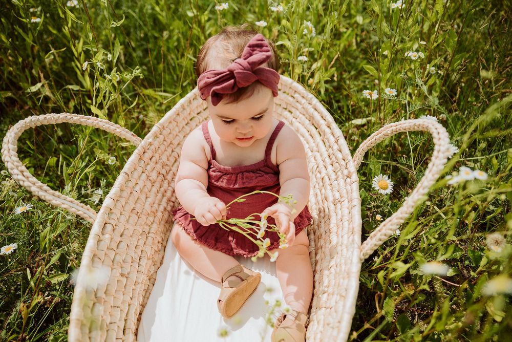 Baby sitting in woven basket surrounded by wildflowers and greenery.