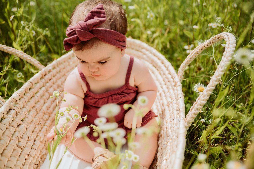 Infant in burgundy outfit sitting in natural wicker basket outdoors.
