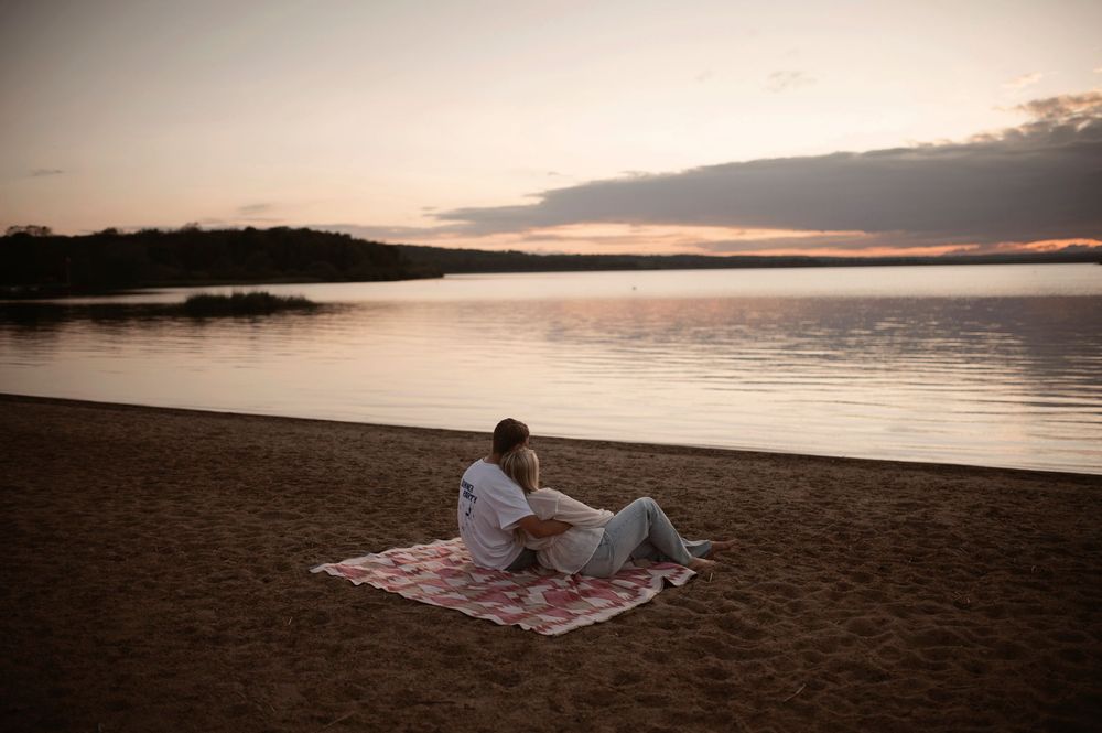 A tranquil beach scene at sunset with figures relaxing on a blanket by the calm water.