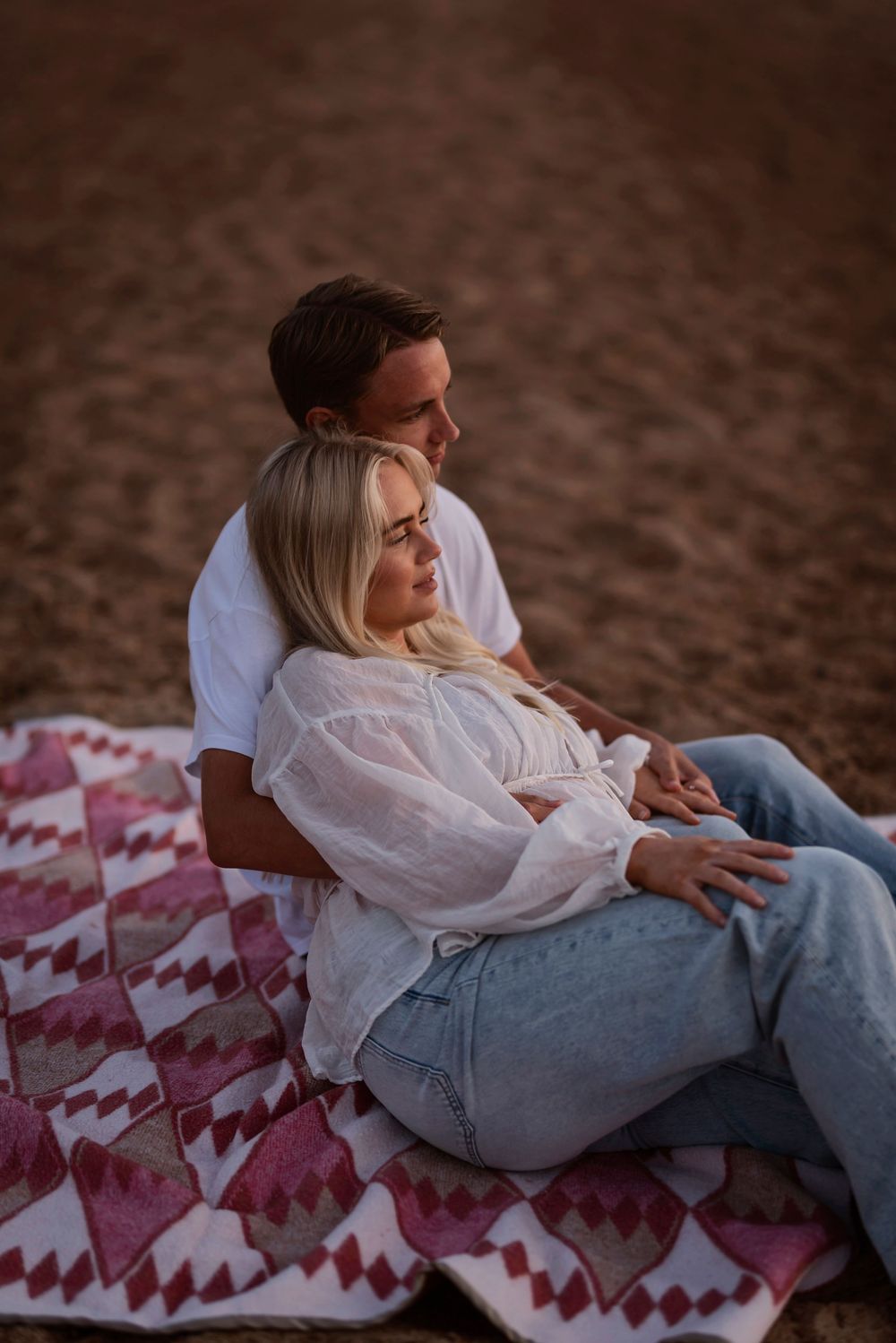 A couple relaxes together on a red and white patterned blanket during a romantic sunset photoshoot.