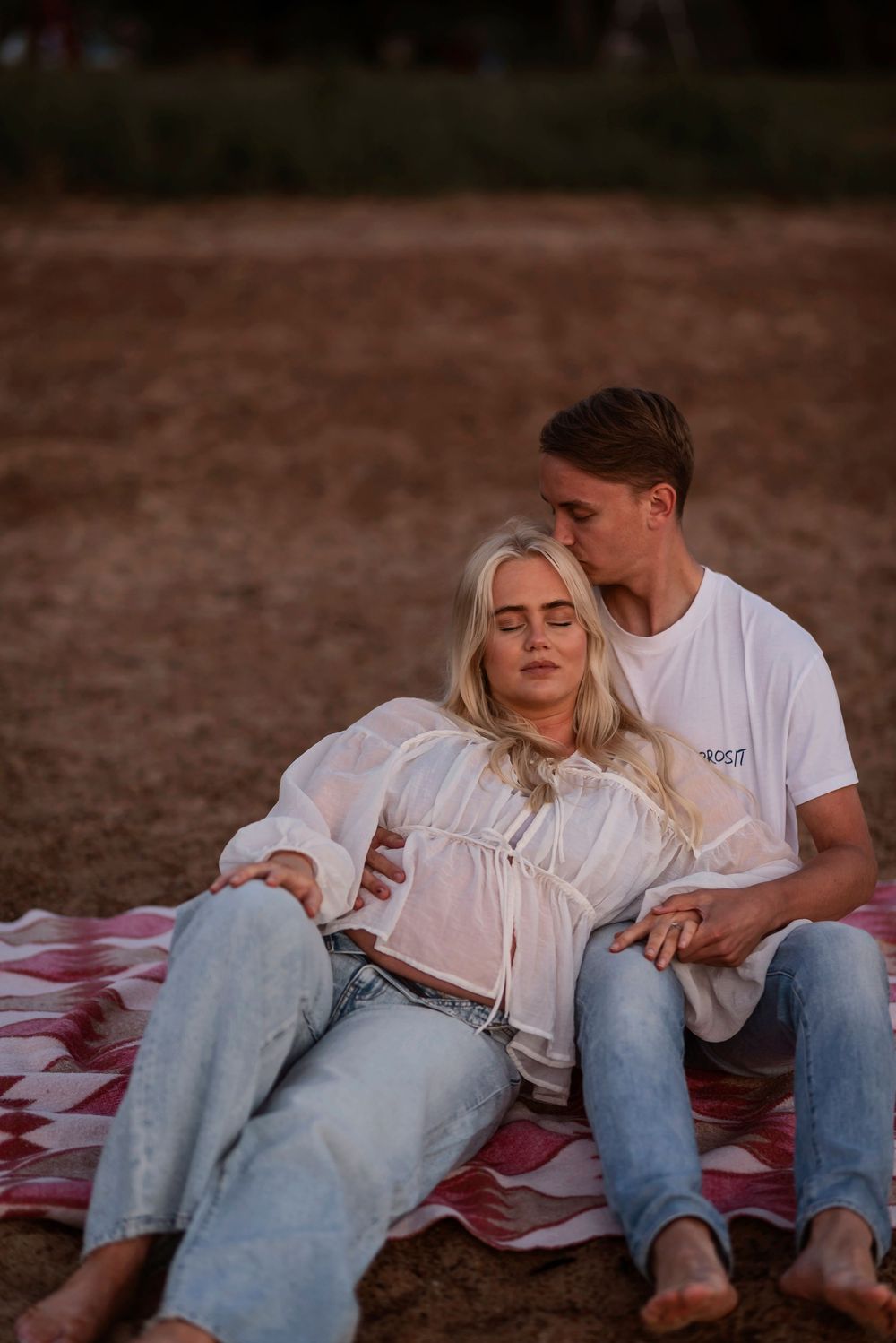 A romantic couple lounges together on a blanket at sunset in casual denim and white outfits.