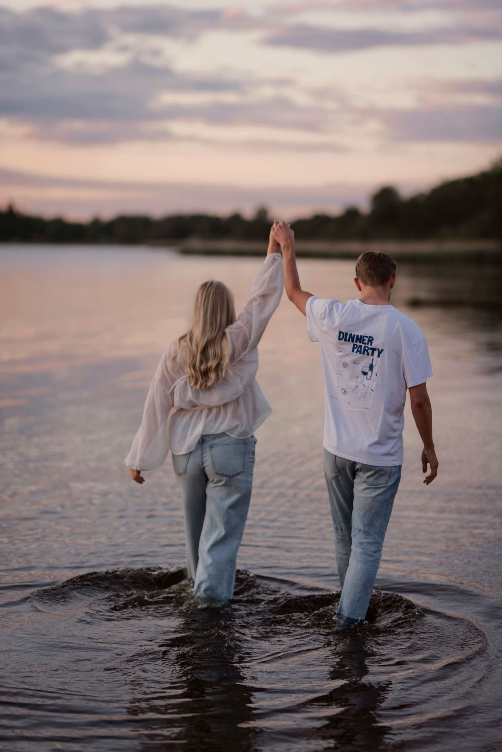 A couple holds hands while wading in water at sunset with cloudy skies above.