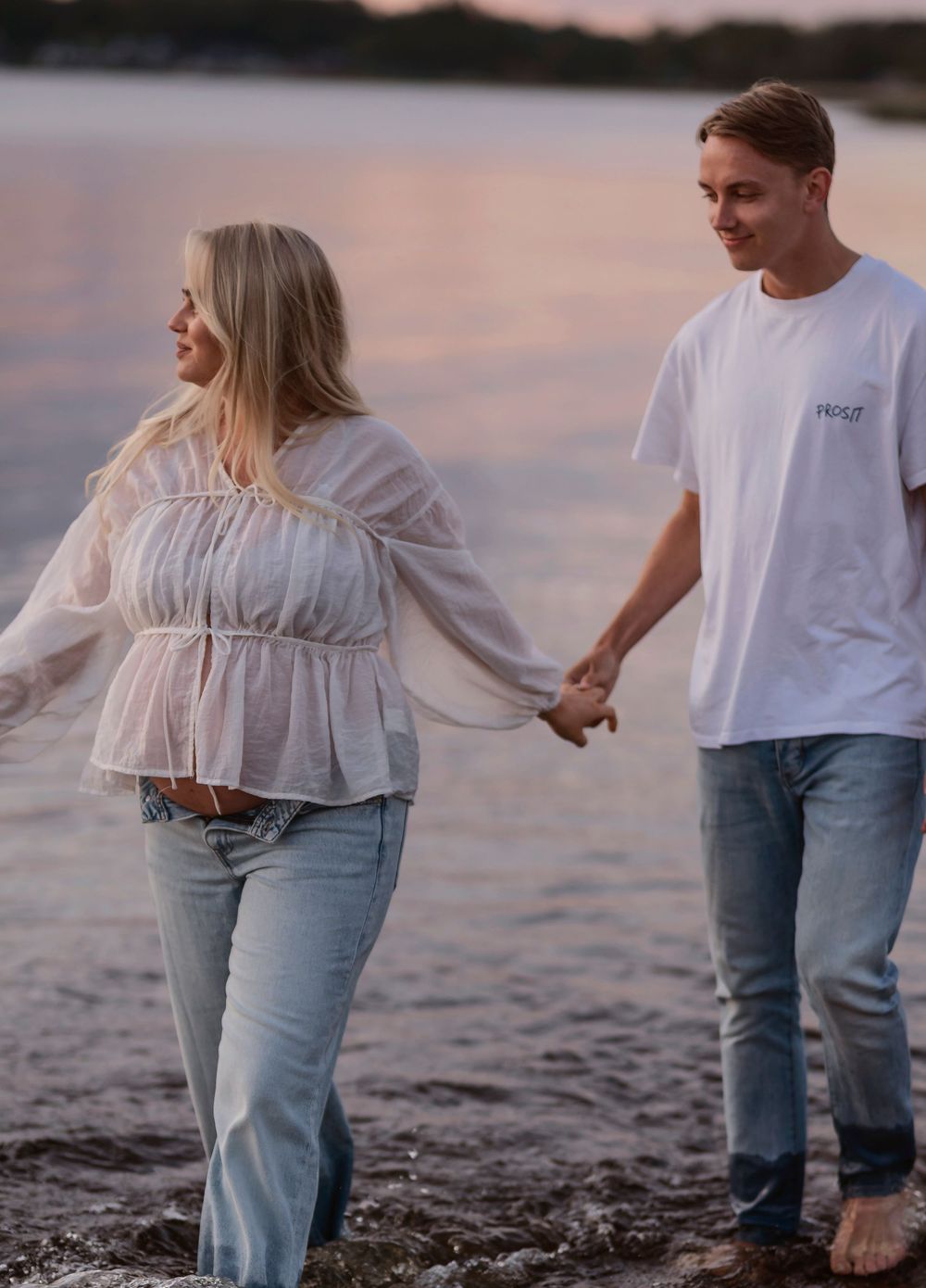A couple holds hands while walking along the beach at sunset wearing casual outfits.