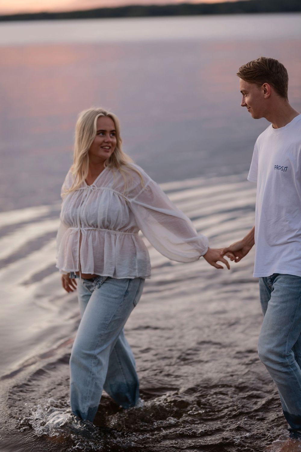 A couple walks along the beach at sunset holding hands while splashing in the shallow water.