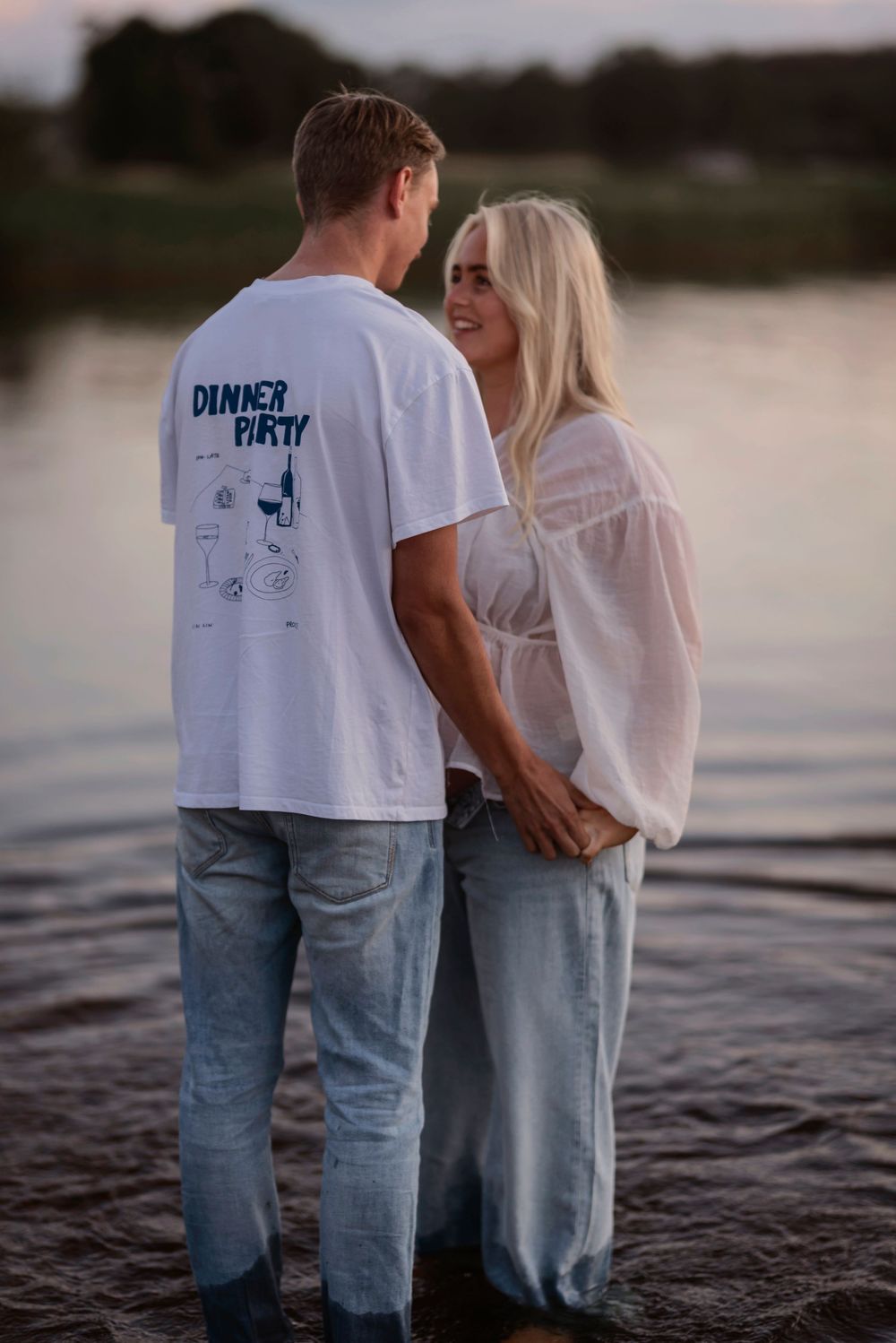 Romantic couple in matching white and denim outfits standing by a lake at sunset.