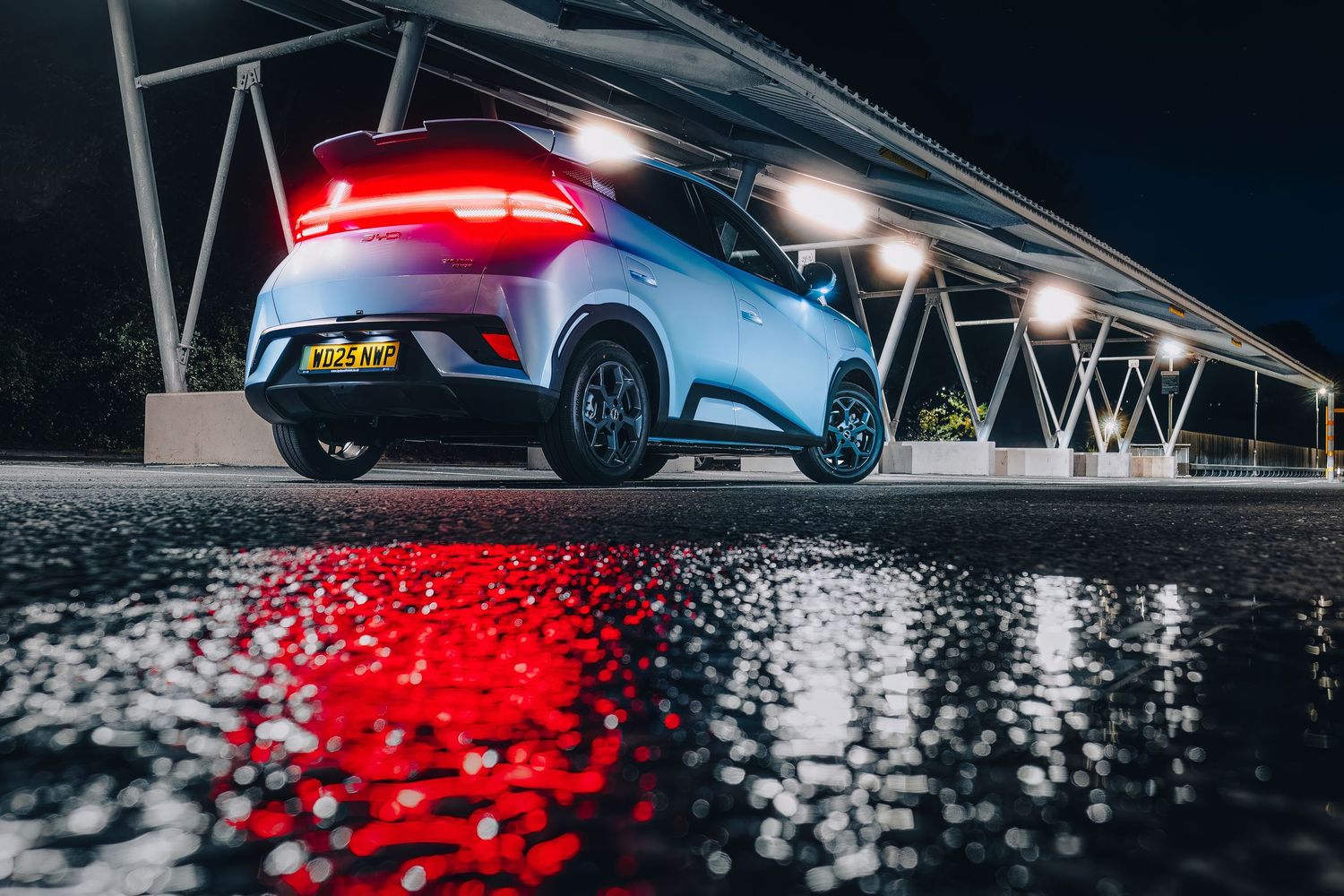 Stylish light blue BYD Dolphin Surf  parked in modern parking lot at night with red light streaks in background.
