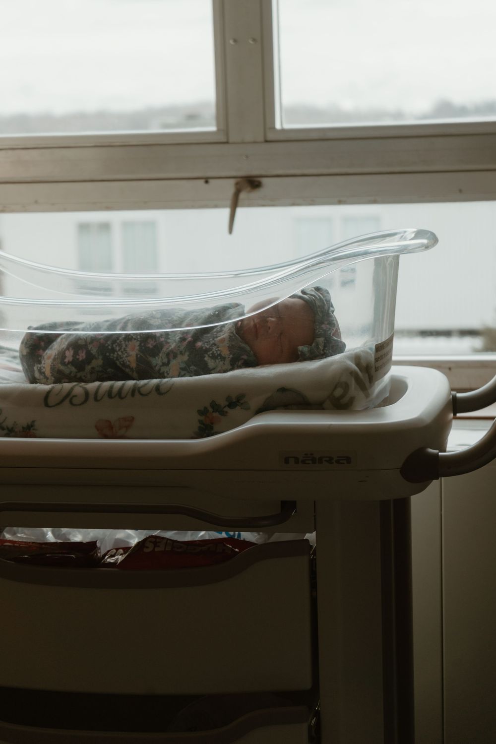 Swaddled newborn baby sleeping peacefully in a clear hospital bassinet by a window.