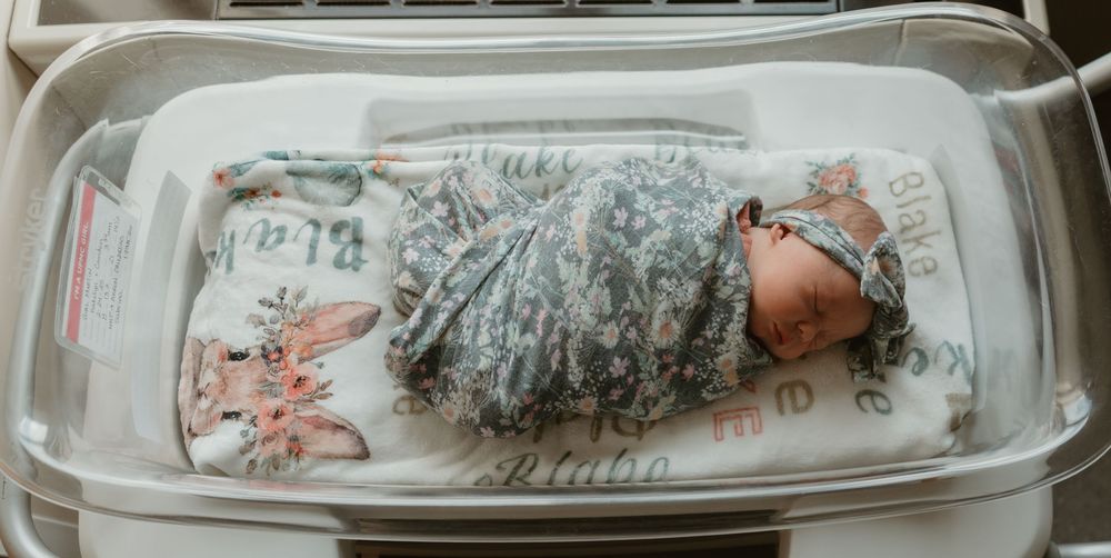 Newborn baby swaddled in a floral blanket with a matching headband sleeping in a hospital bassinet.