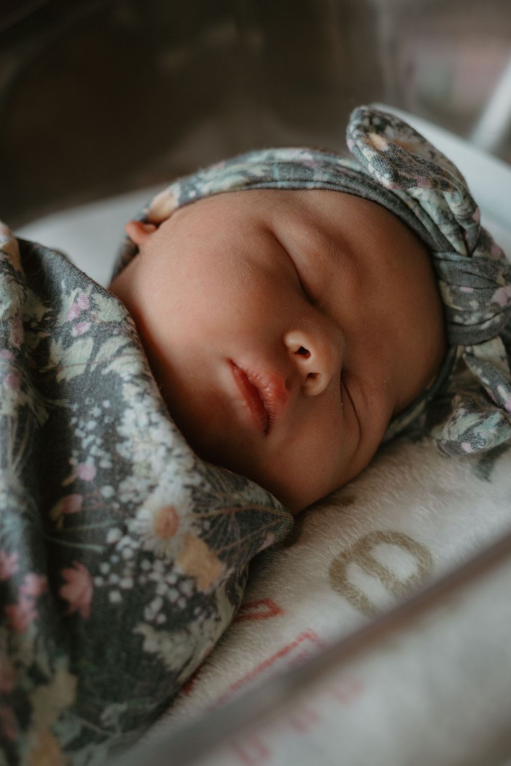 Sleeping newborn baby wrapped in floral swaddle blanket wearing matching bow headband in hospital bassinet.